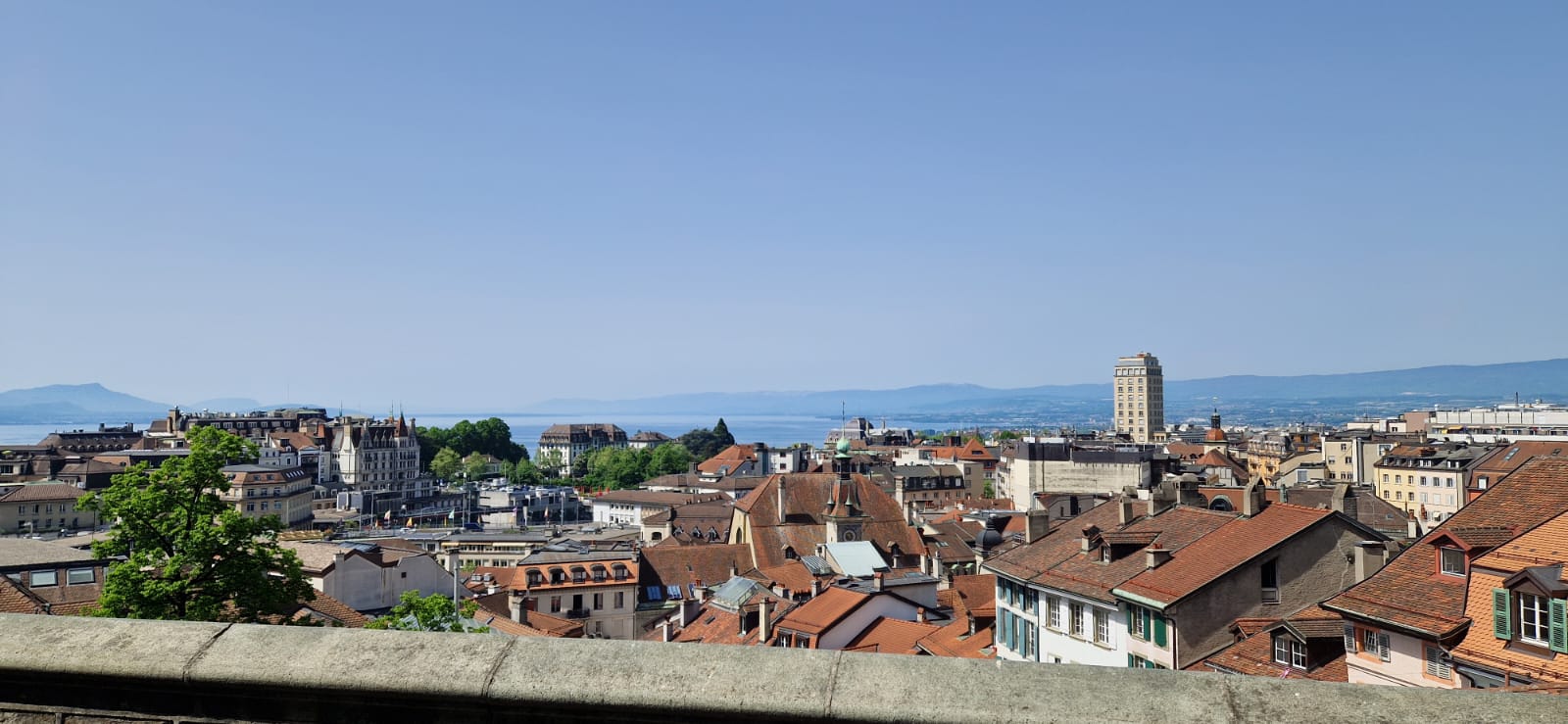 Cityscape with red-tiled roofs, tall building, water, mountains, and clear blue sky in the background.