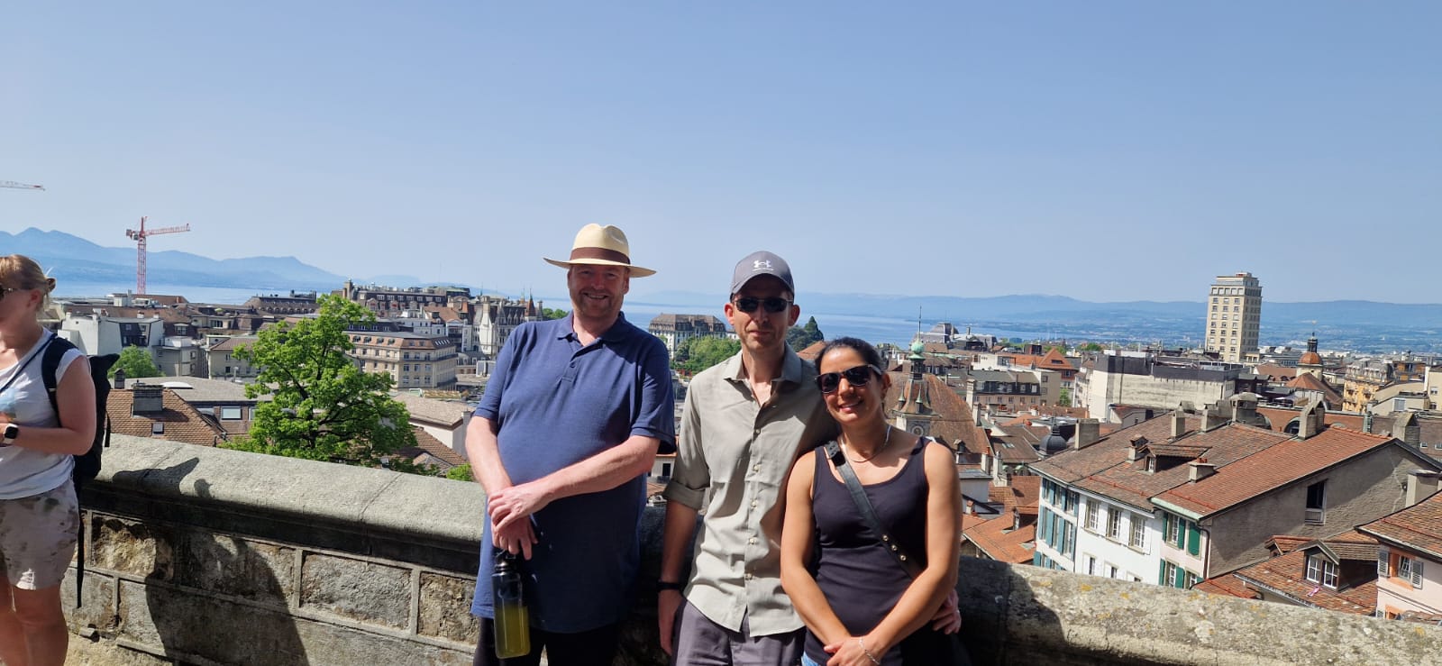 Three people on a stone balcony with a cityscape, rooftops, and mountains under a clear sky.