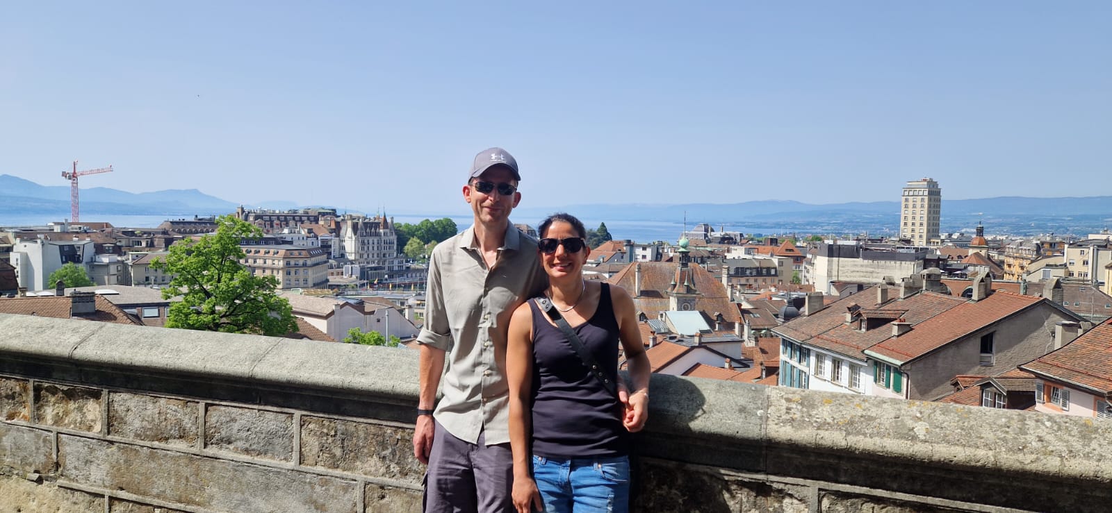 Two people on a terrace overlook a city with red roofs, water, and mountains in the background.