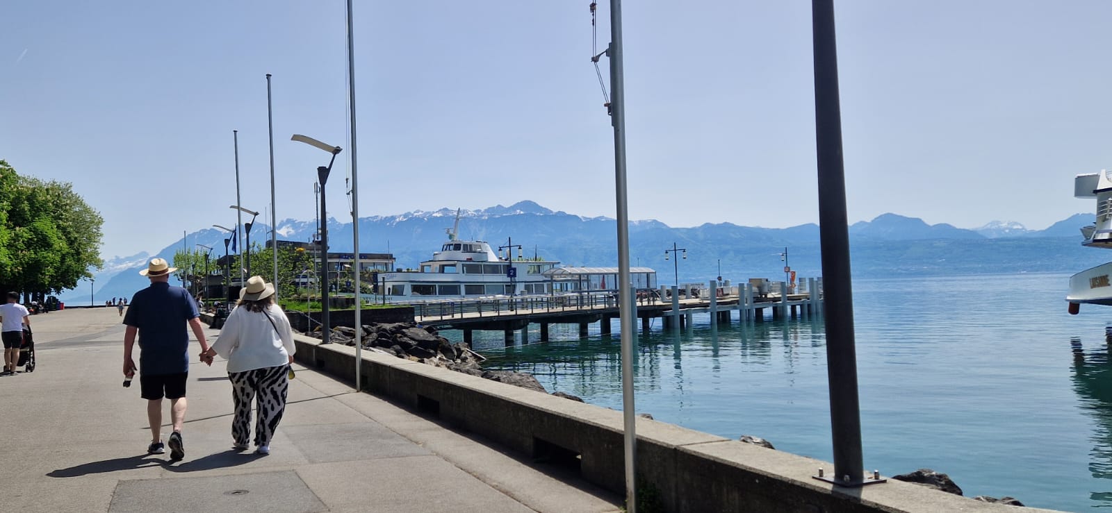 Couple walks on promenade near water and dock, mountains in distance, clear sunny sky.