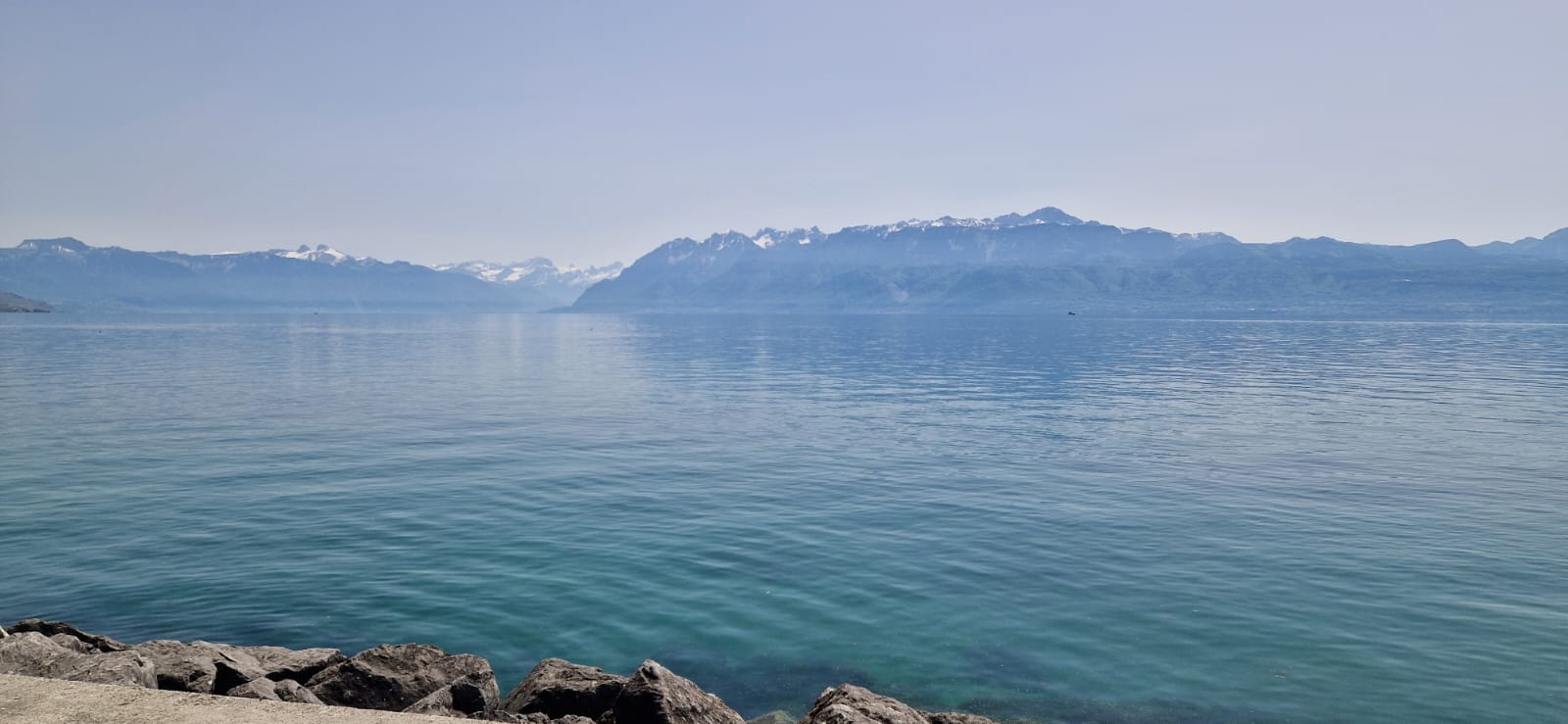 Calm water with gentle waves, rocky shore, snow-capped mountains, clear blue sky.