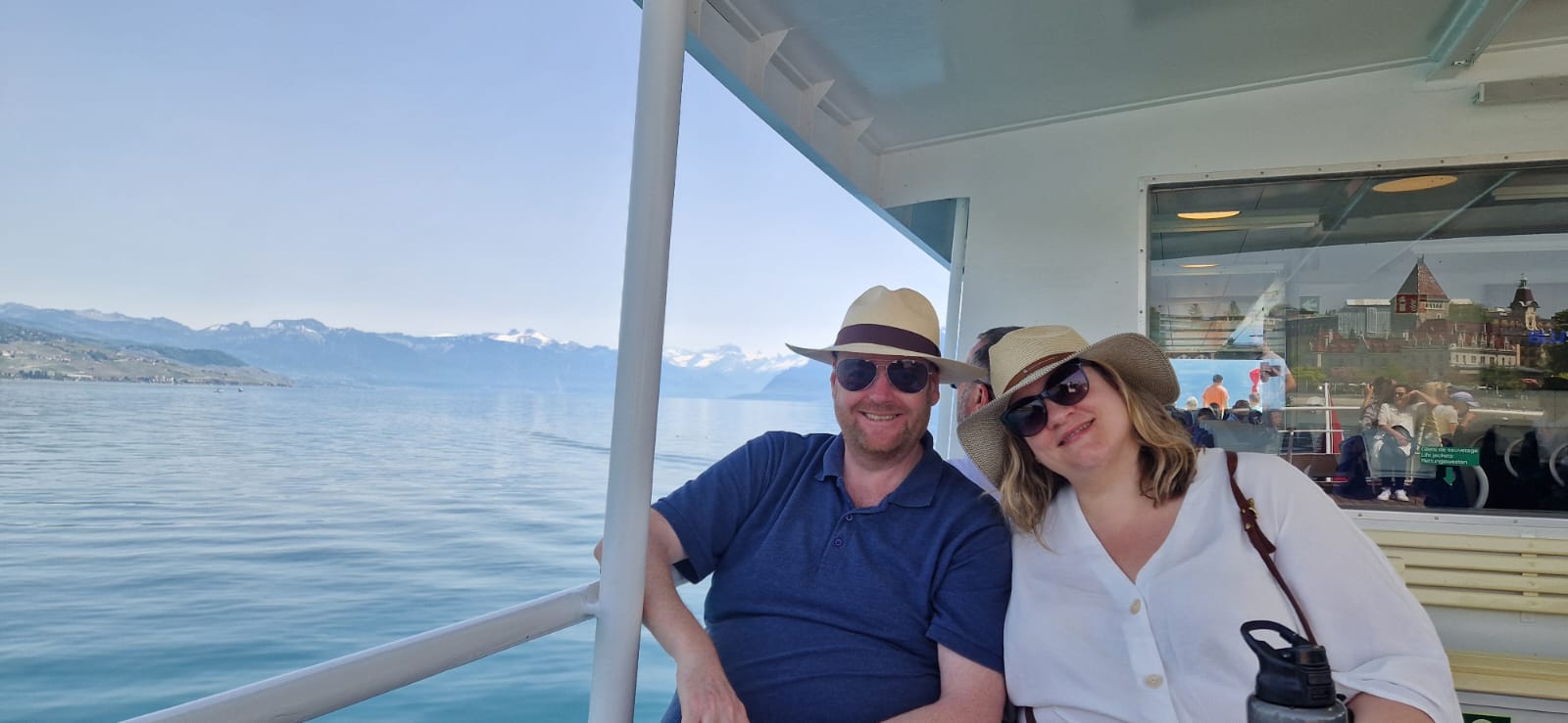 Man and woman on a boat wearing sunglasses and straw hats, with water and mountains behind.