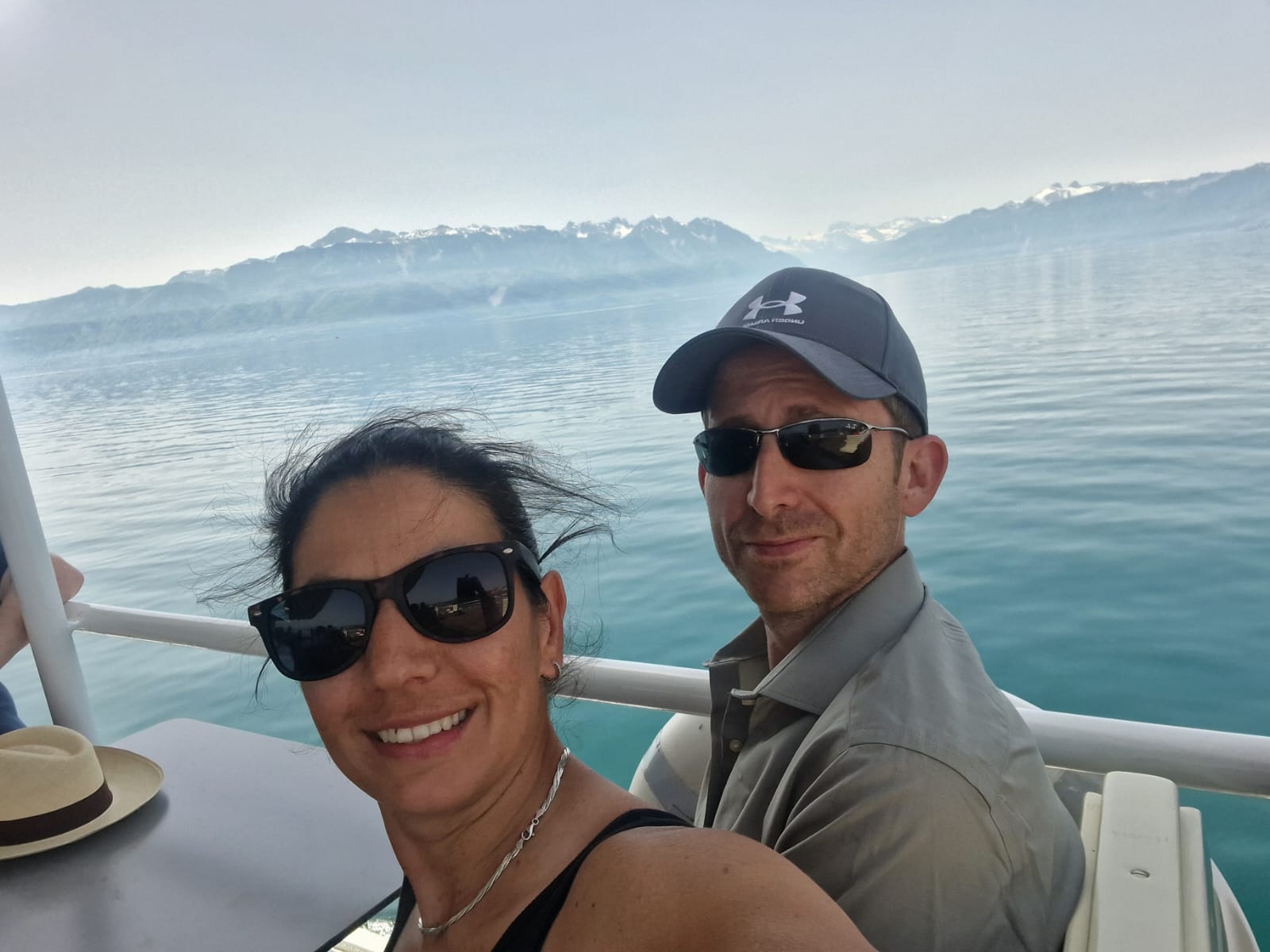 Two people taking a selfie on a boat with water, mountains, clear sky; wearing sunglasses, cap.