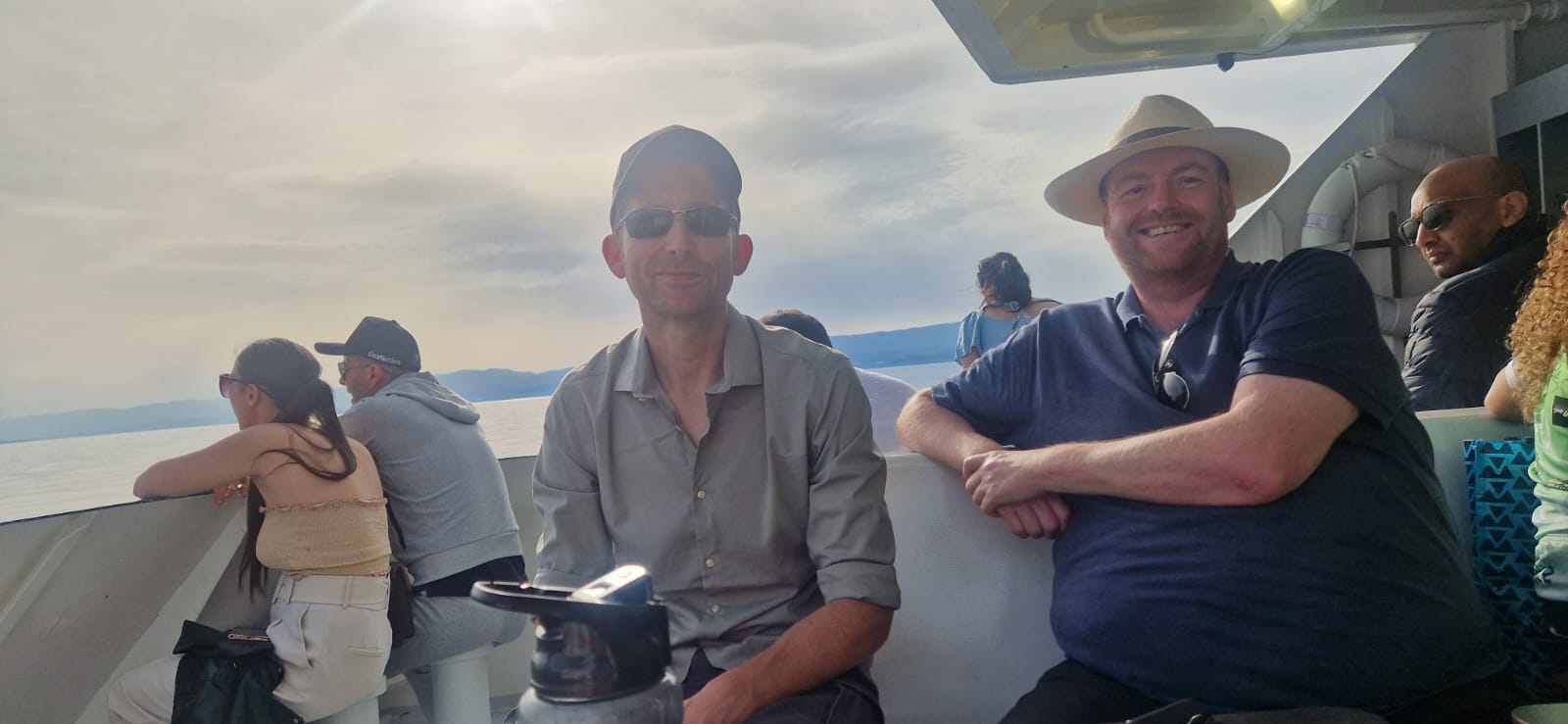 Men sitting on a boat, one in a baseball cap and sunglasses, the other in a straw hat.