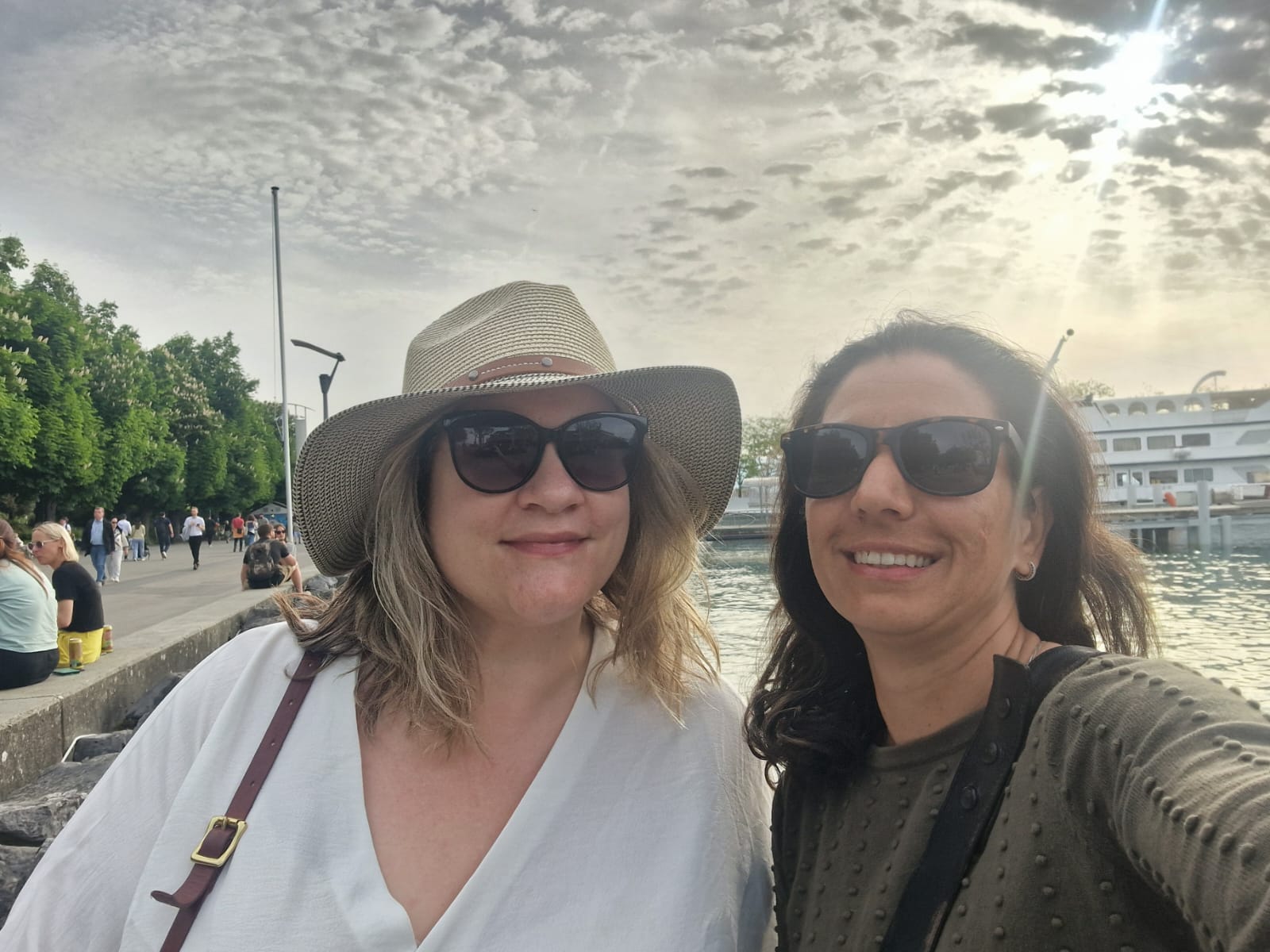 Two people outdoors near waterfront, wearing sunglasses; boat and sunny sky in background.