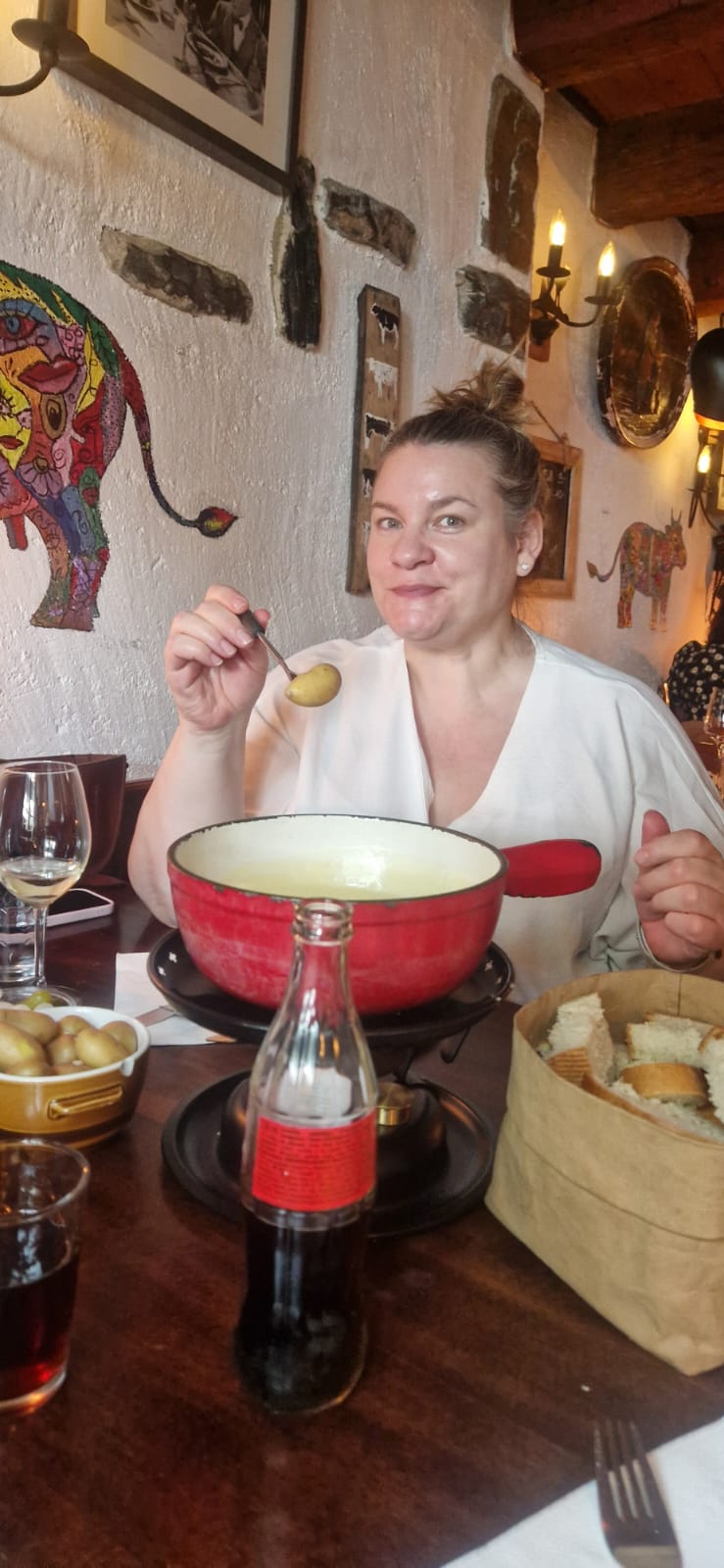 Person enjoying cheese fondue at a table with bread, potatoes, soda, and colorful wall art.