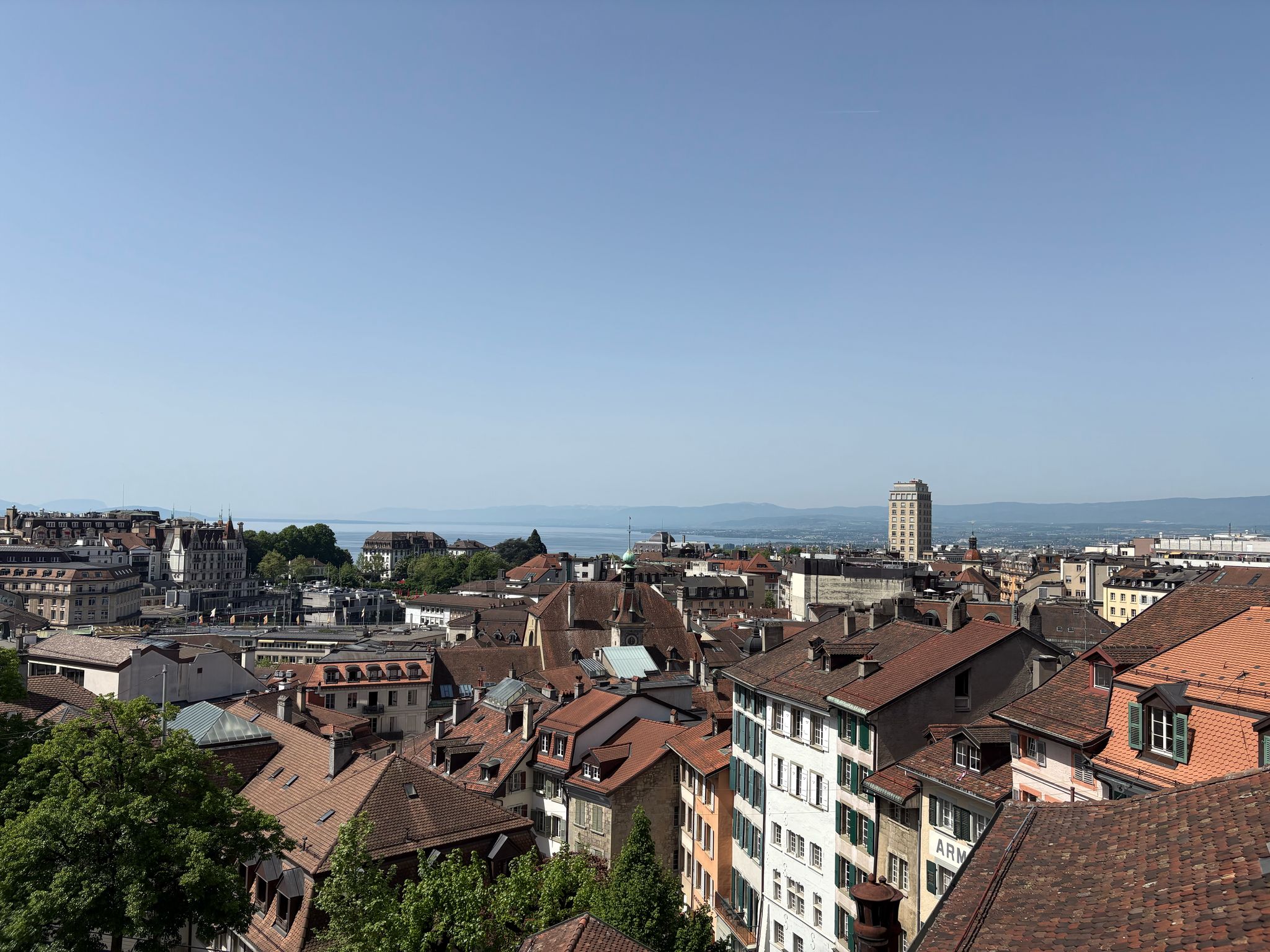 Cityscape with red-tiled roofs, a prominent tower, distant water, and hills under a clear sky.