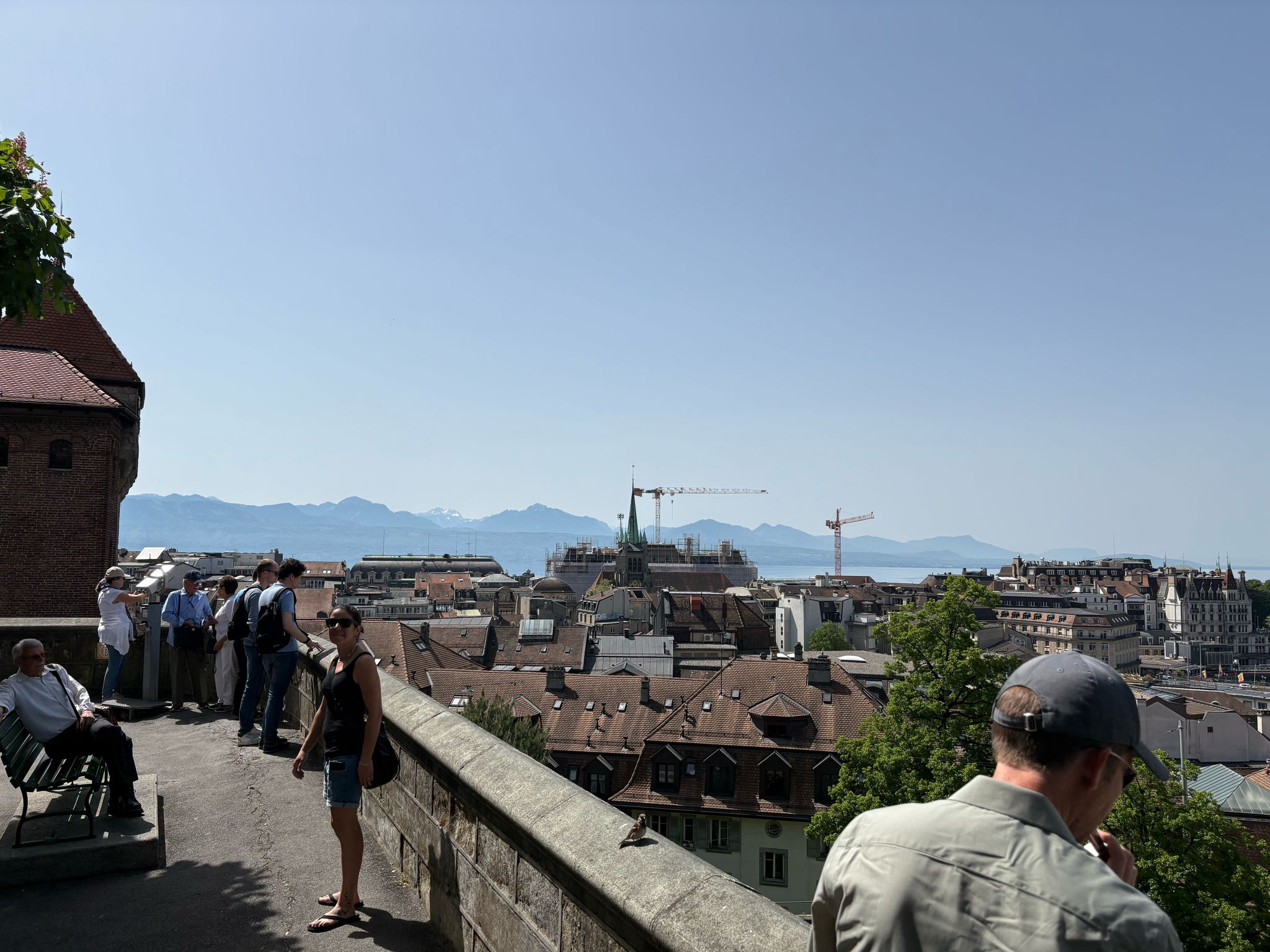 People on a walkway overlooking cityscape with cranes, mountains, clear sky. Some take photos.