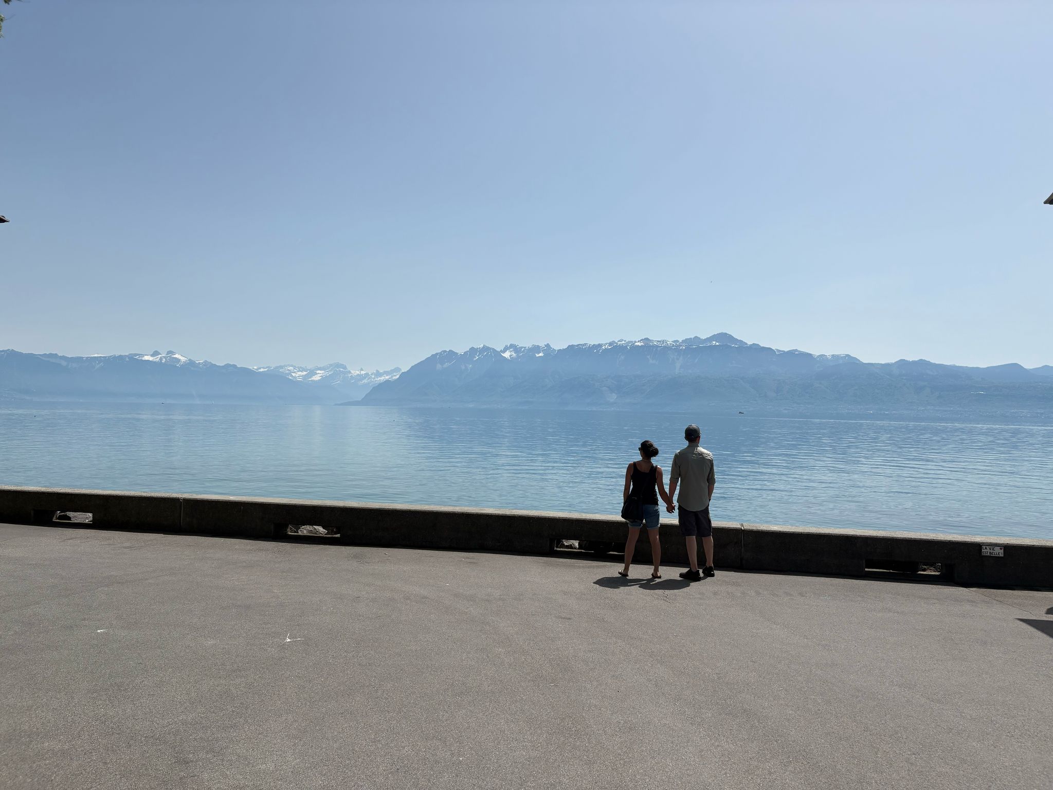 Couple on paved walkway by water, holding hands, viewing snow-capped mountains, clear sky.