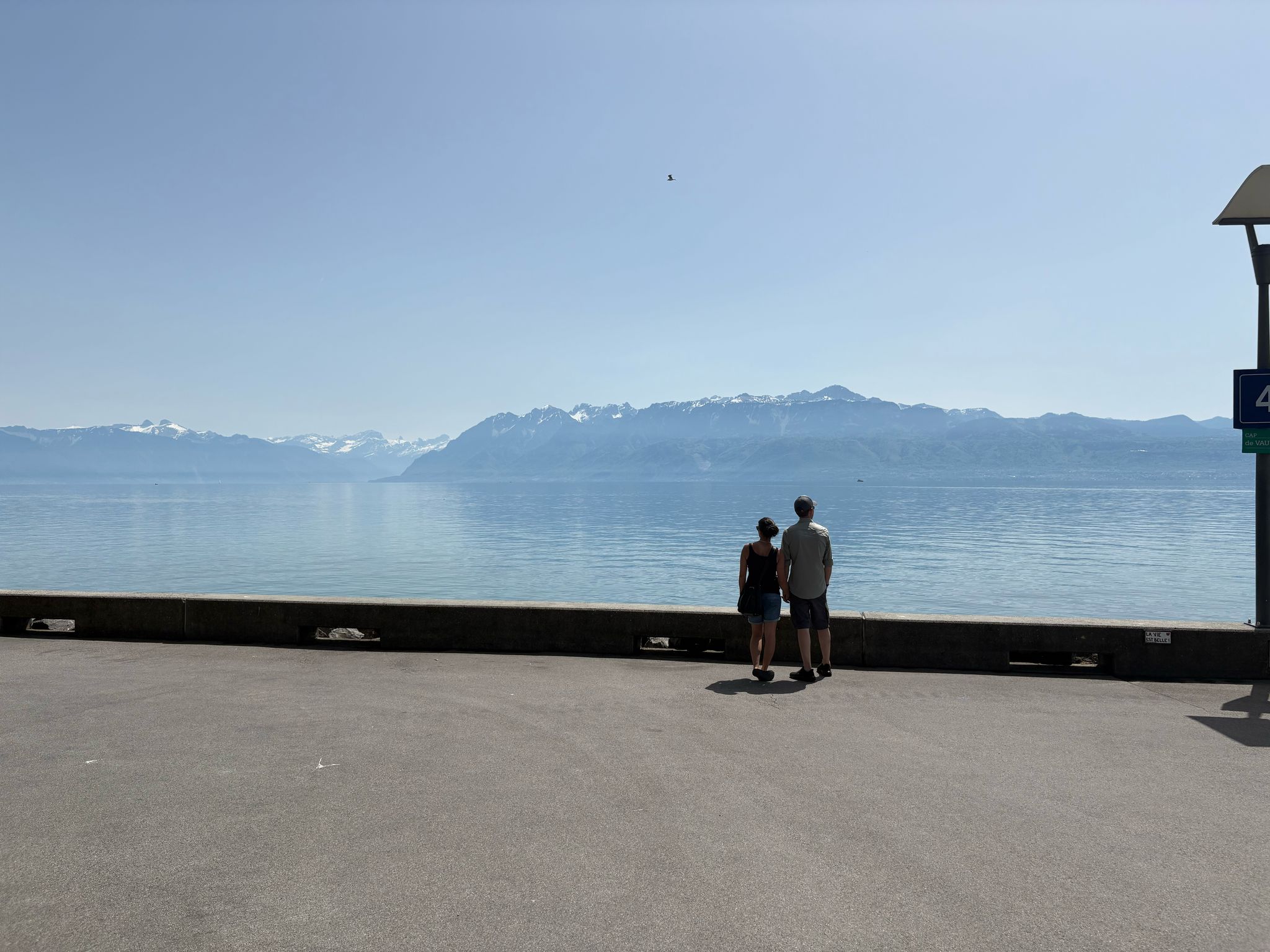 Couple on paved area overlooking calm water and snow-capped mountains under clear sky.