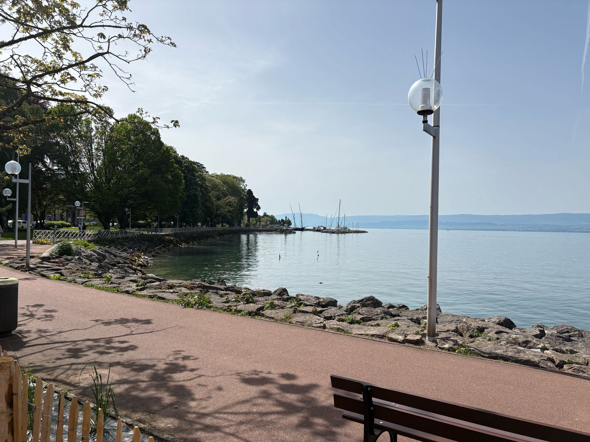 Lakeside Walkway with Sailboats Lakeside walkway with trees, lampposts, calm water, distant sailboats, and a bench under clear sky.
