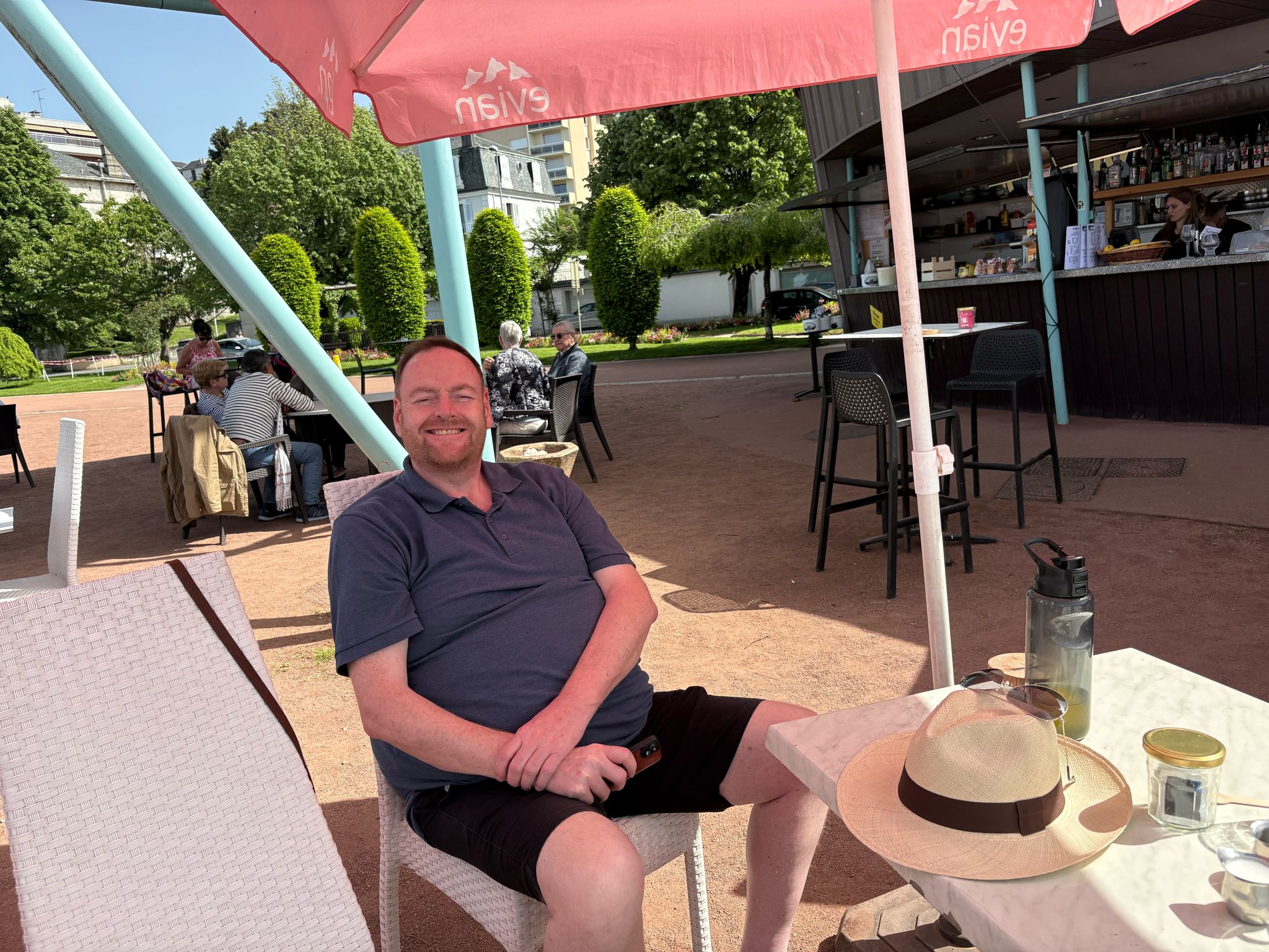 Man relaxing at outdoor cafe under pink Evian umbrella, table with hat and bottle nearby.