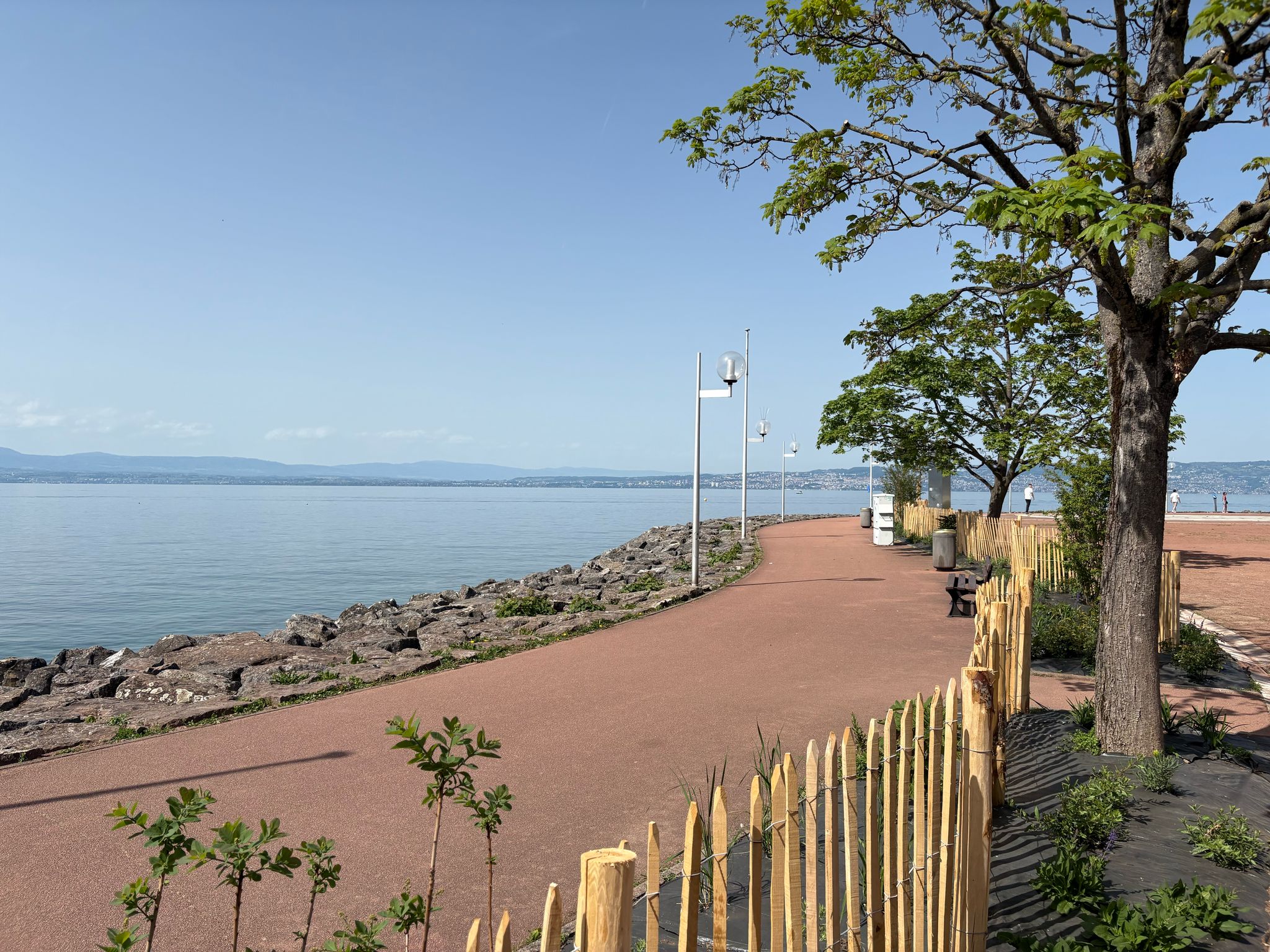 Lakeside Walkway with Mountain Views Lakeside walkway with red pavement, wooden fencing, trees, rocks, and distant mountains.