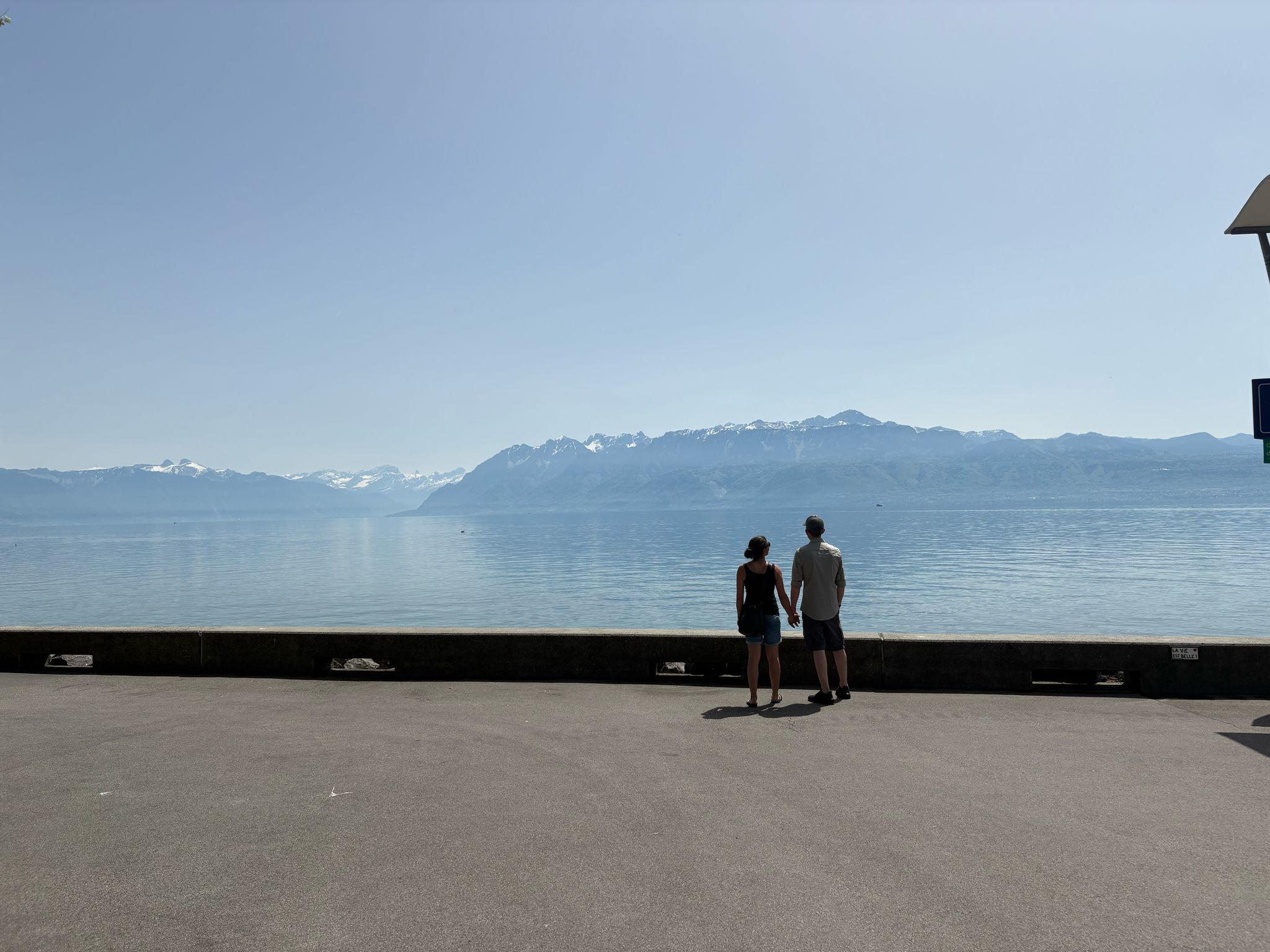 Couple on walkway facing calm water and distant snow-capped mountains under clear sky.