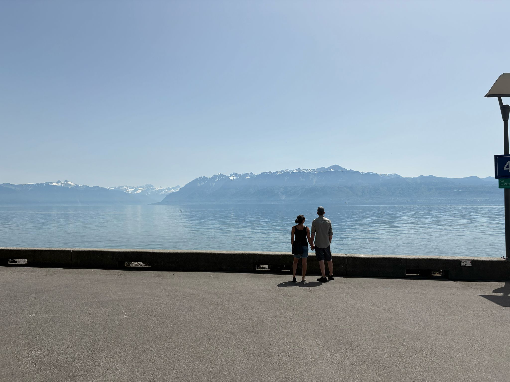 Man and woman stand near calm water, looking at distant mountains under clear blue sky.