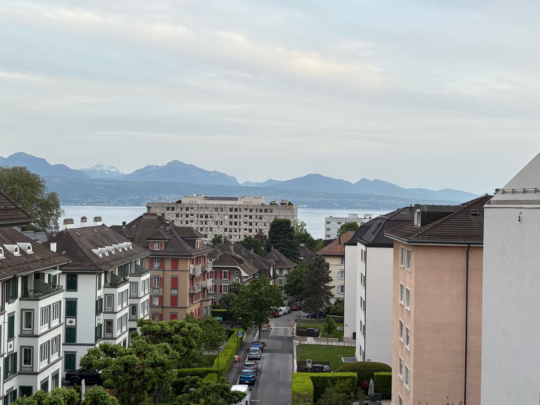 Residential area with mixed architecture, tree-lined streets, lake, and mountains under cloudy sky.