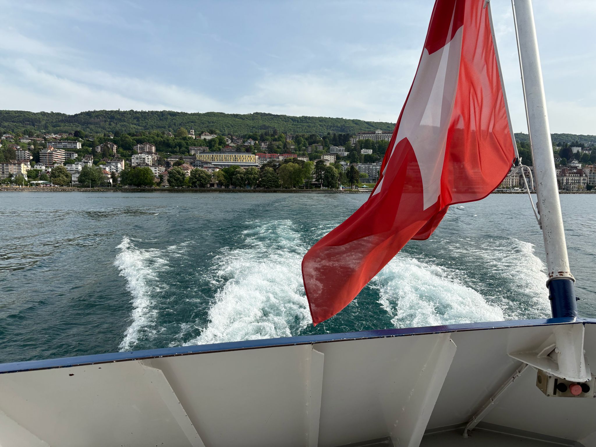 Red flag with white cross on a boat, scenic shoreline with buildings, green hills, cloudy sky.