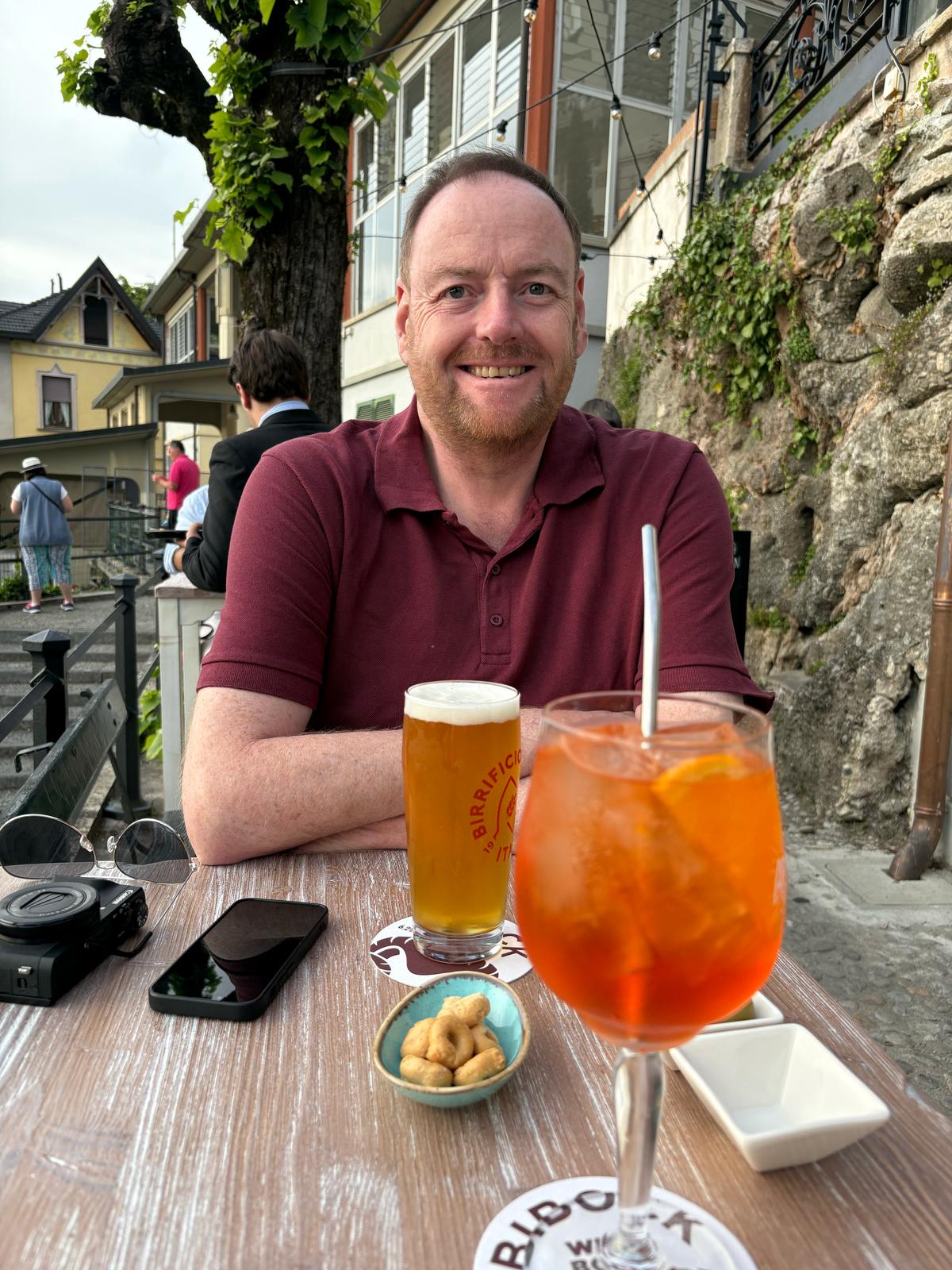 Man sitting at outdoor table with beer, cocktail, snacks, camera, and phone; street background.