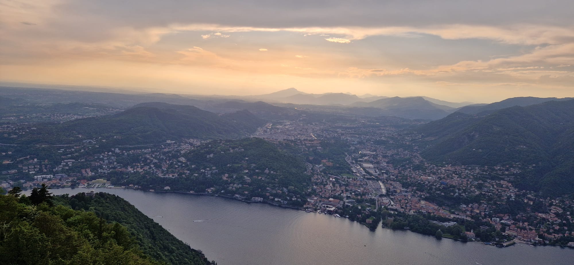 Panoramic view of sunset over lake, forests, urban area, and silhouetted mountains.