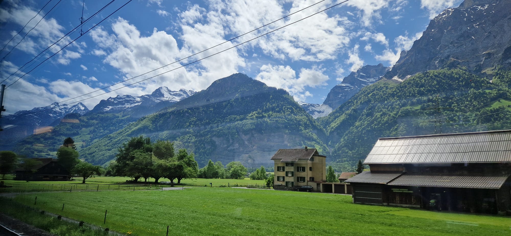 Green fields, trees, farmhouse, and building; snow-capped mountains and power lines against cloudy sky.