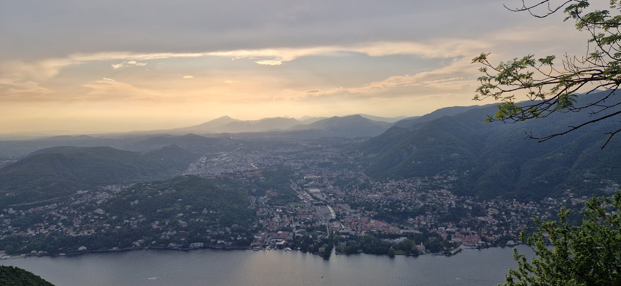 Mountainous Lakeview Panorama Panoramic view of a lake, city, mountains, cloudy sky, and tree branches on the right.
