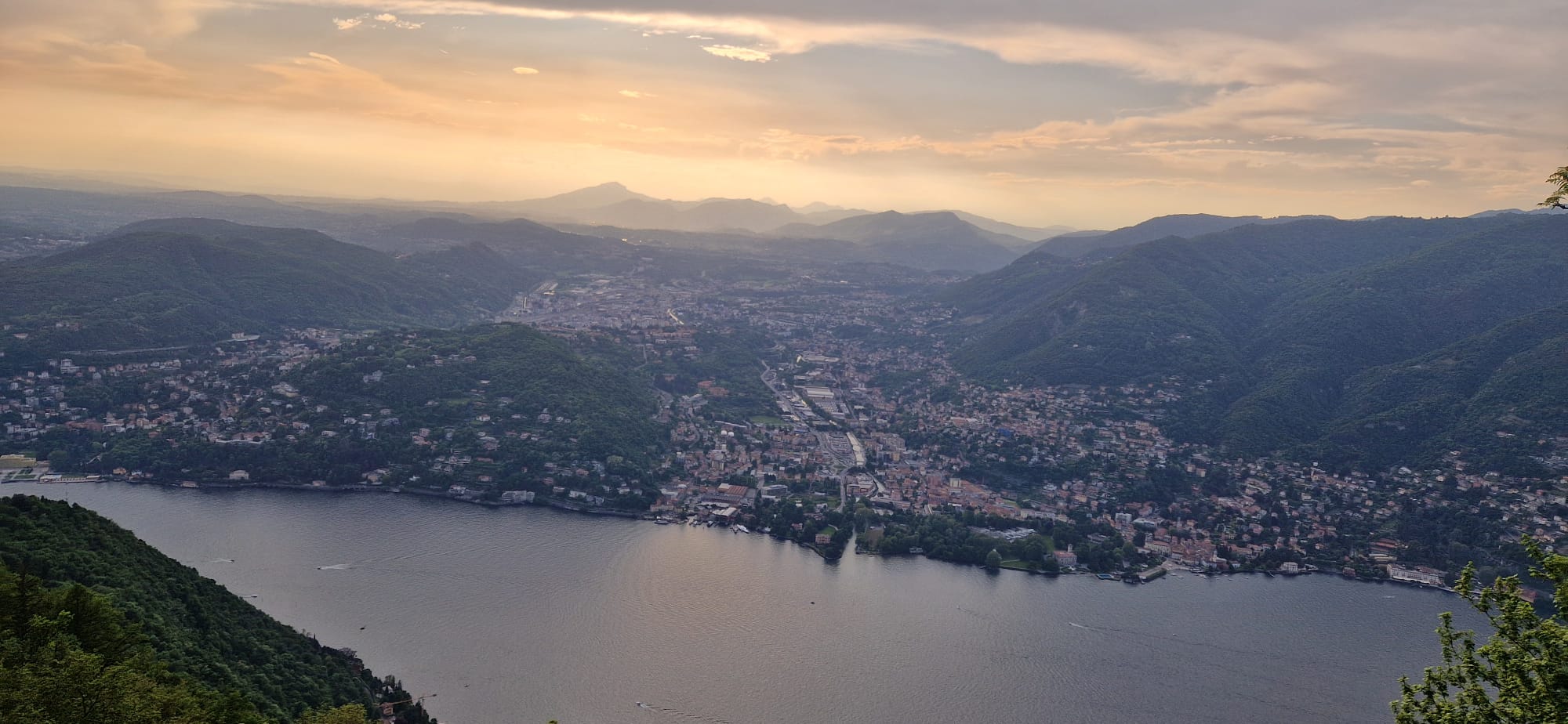 Scenic view of a town by a large lake, surrounded by hills under a cloudy, warm-lit sky.