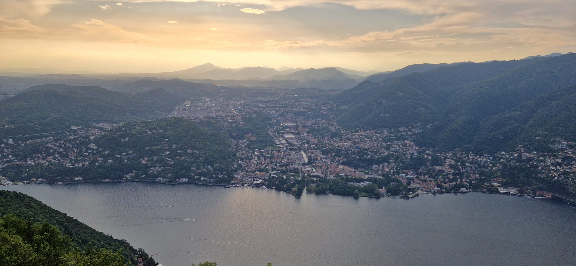 Panoramic view of a serene lake, lush hills, and a town under a sunset sky.