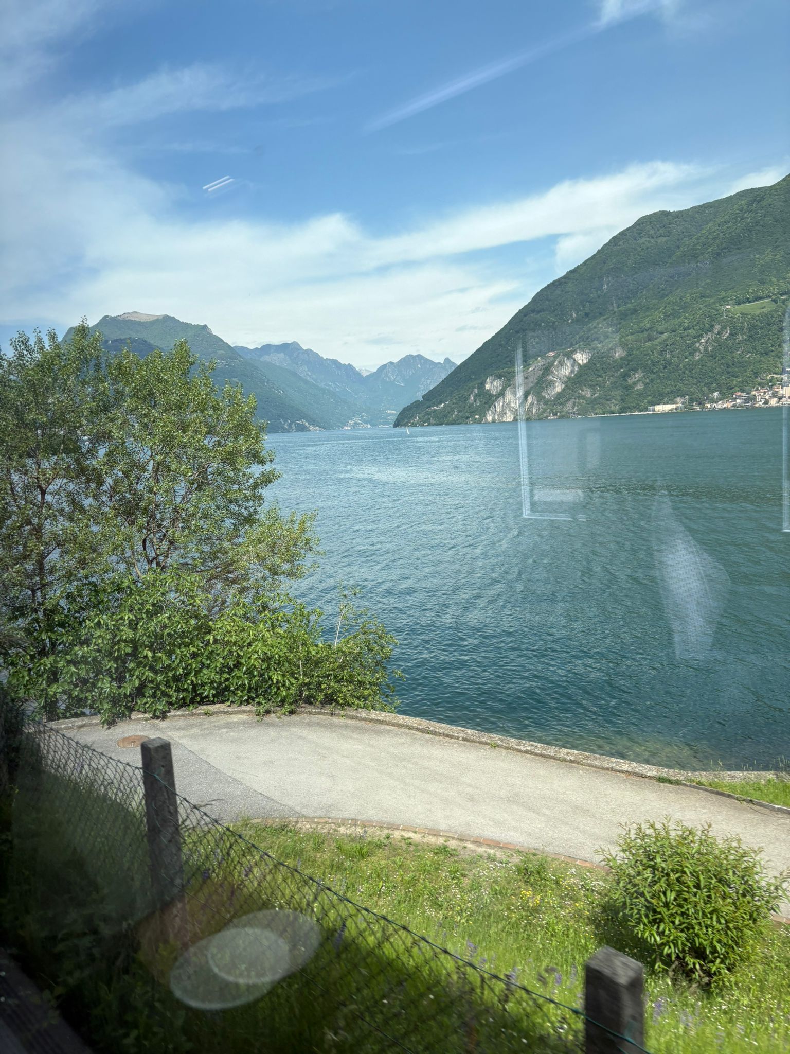 Scenic lake view with green hills, clear sky, path, greenery, and distant mountains.