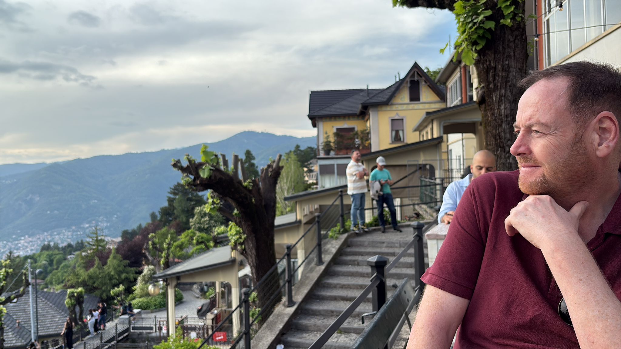 Person in maroon shirt sitting outdoors, mountains and buildings in background, cloudy sky.