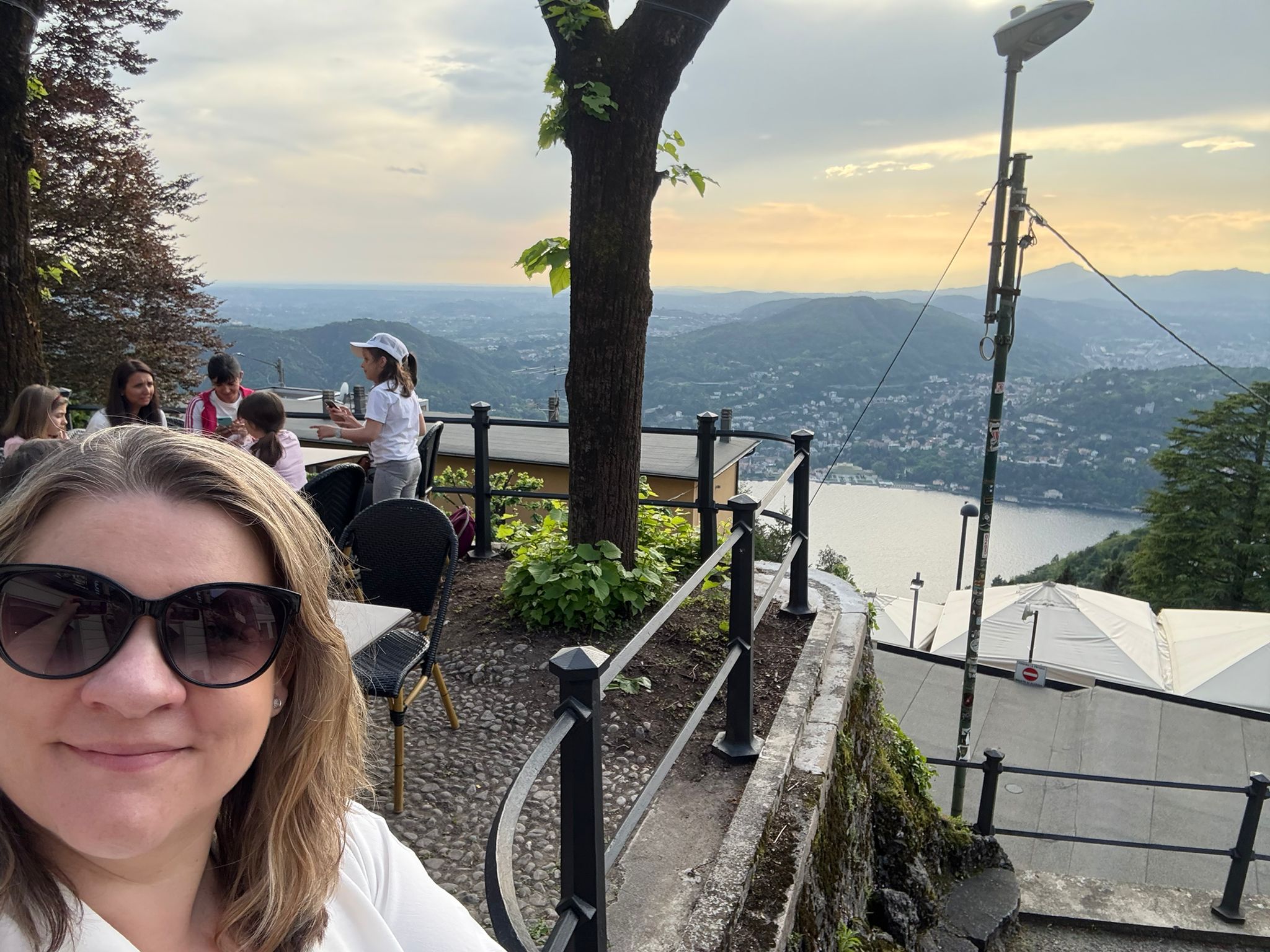 Scenic Terrace Selfie Woman on terrace taking selfie; mountains, water, city view; people seated at table nearby.