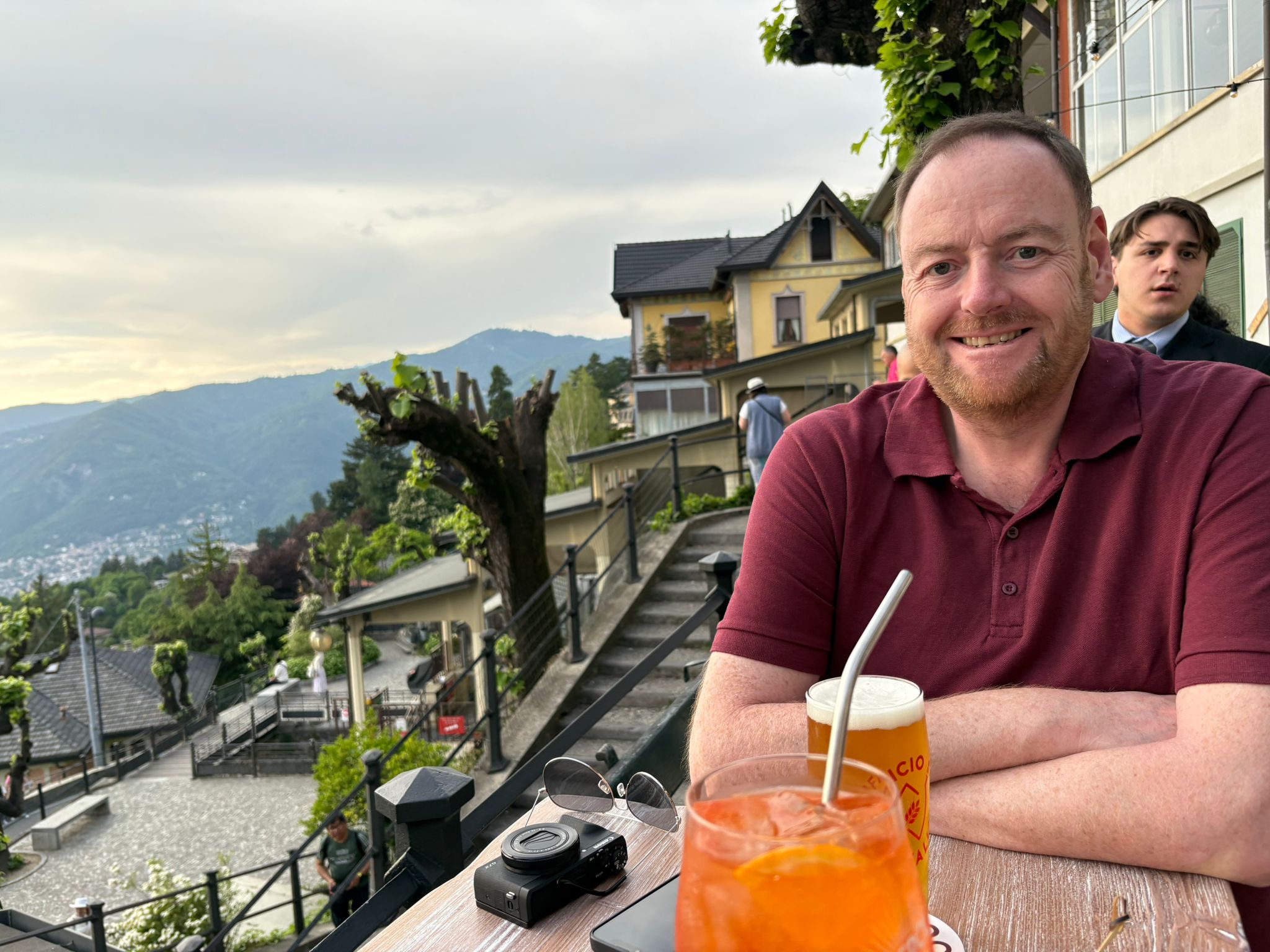 Person enjoying drinks at an outdoor table with scenic view, camera and sunglasses on the table.