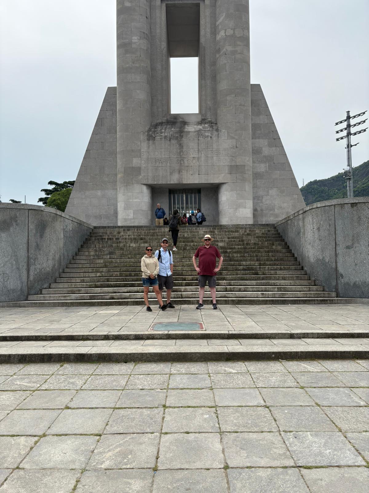 Large stone monument with steps, people standing and sitting, overcast sky, minimal vegetation.