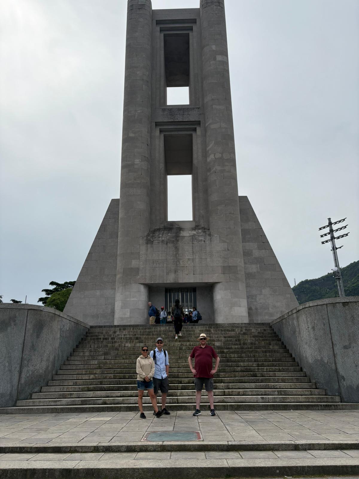 Tall gray monument with two pillars, people pose on steps, overcast sky, greenery in background.