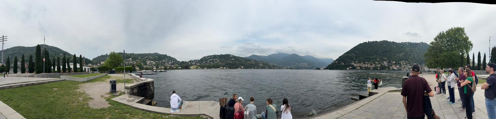 Lakeside Walkway Gathering Panoramic lakeside view with people on a walkway, water, hills, and overcast sky.