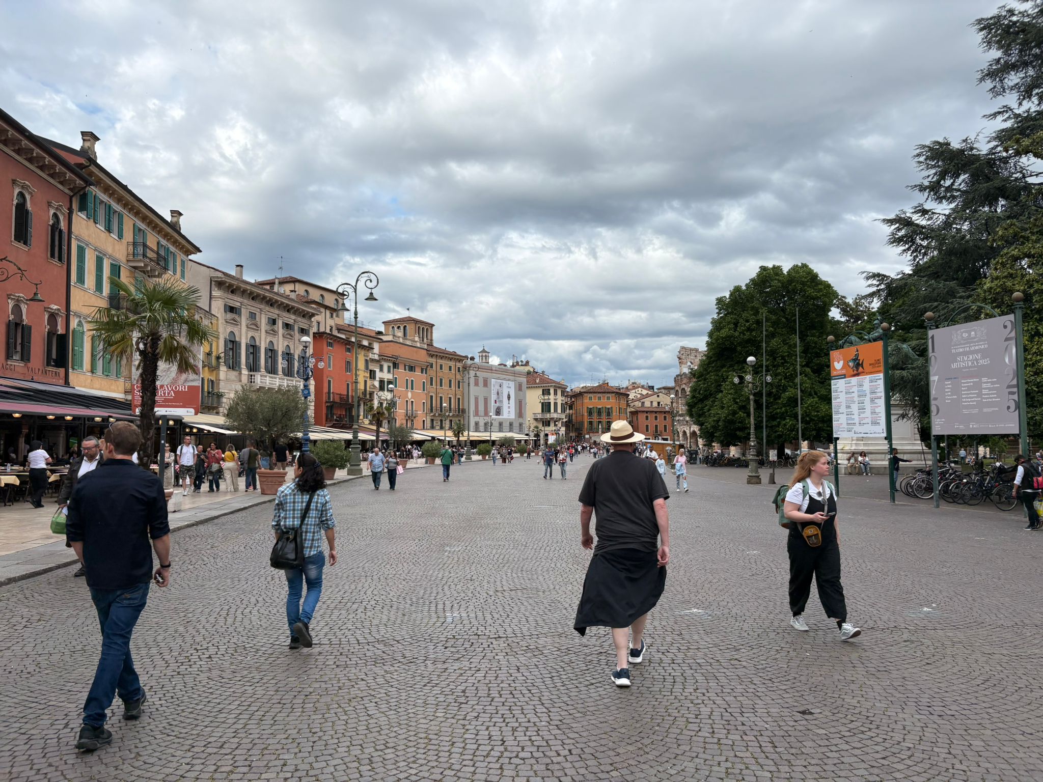 Outdoor plaza with cobblestone, colorful buildings, people, trees, signs, bicycles, cloudy sky.