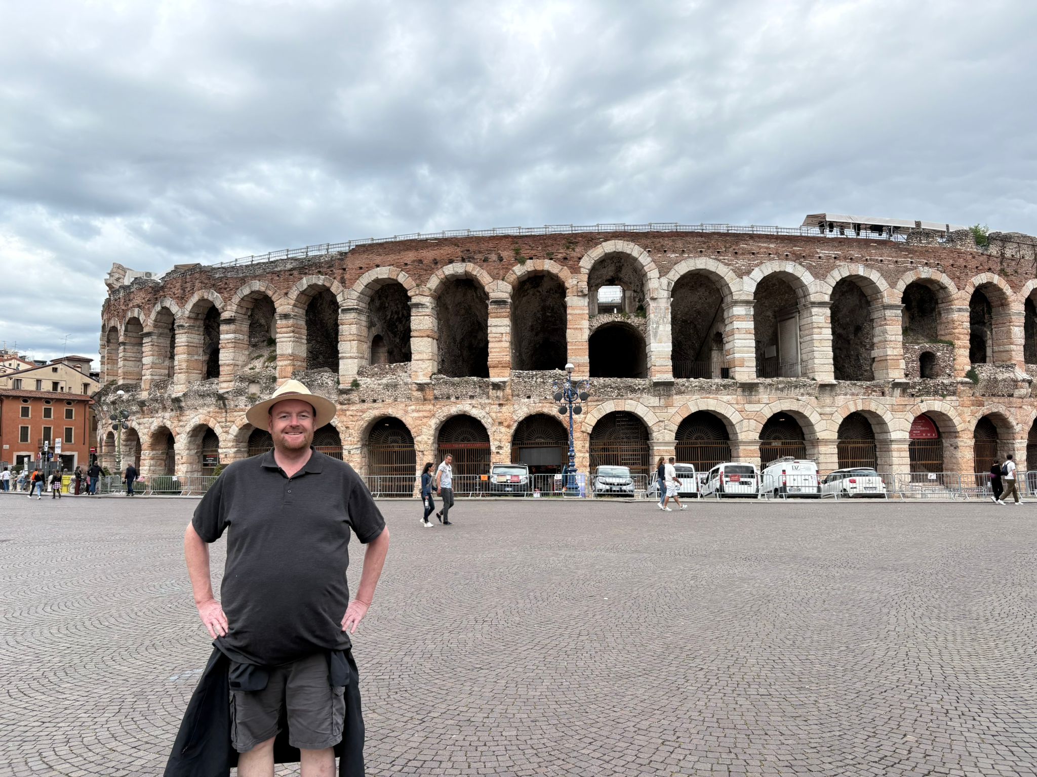 Man wearing a hat in front of an ancient stone amphitheater with arched openings, cloudy sky.