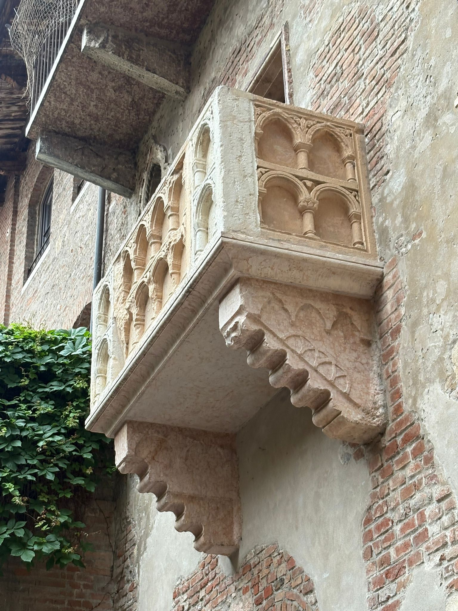 Stone balcony with arches and corbels on a brick wall, vines growing nearby.