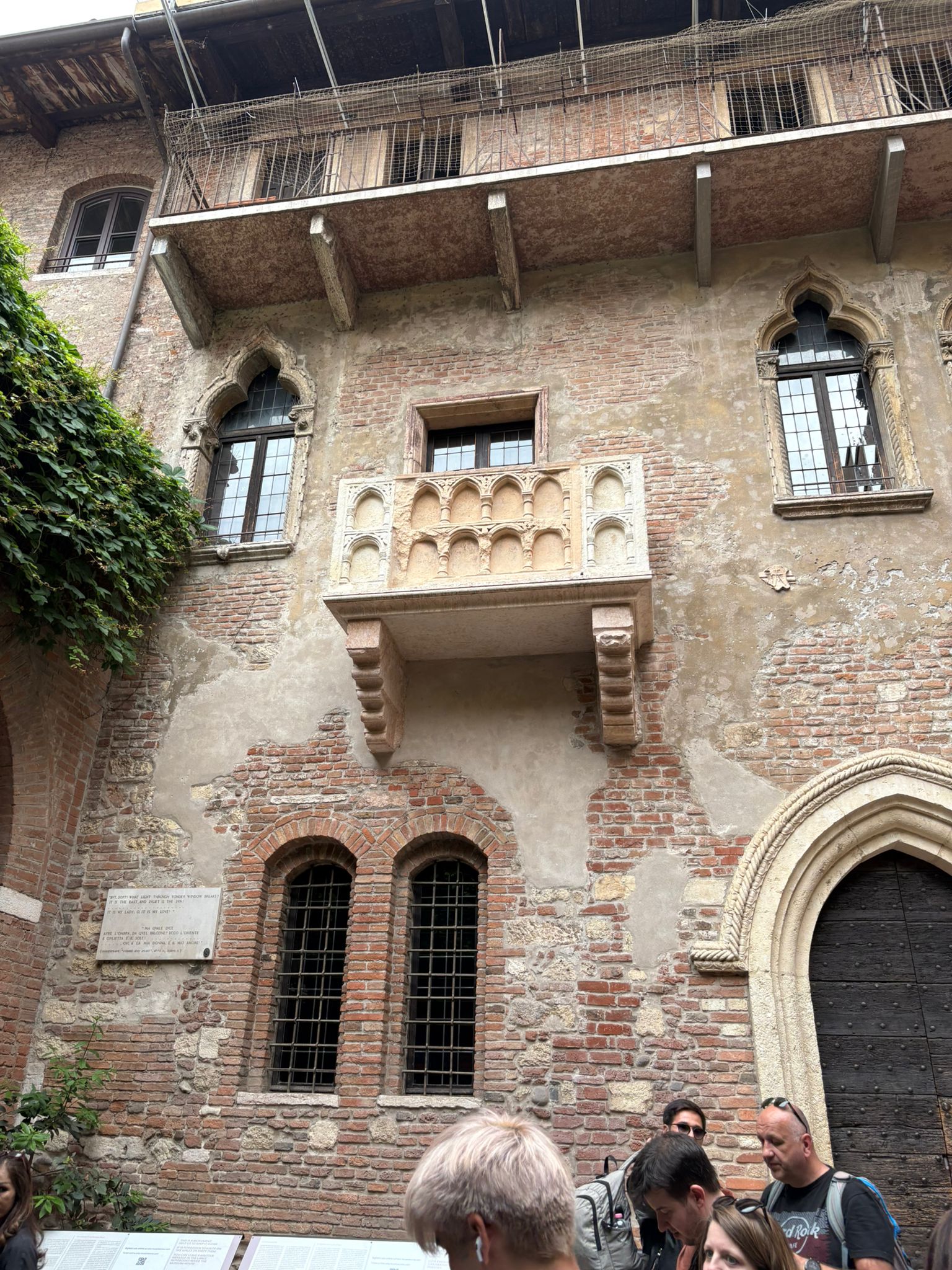 Historic brick building with stone balcony and ornamental windows; people observing below.