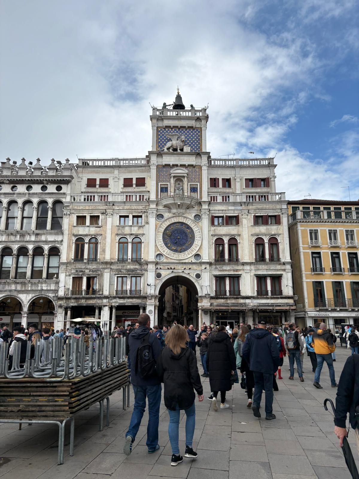 Busy plaza with people, ornate historical building with clock and statue, partly cloudy sky.