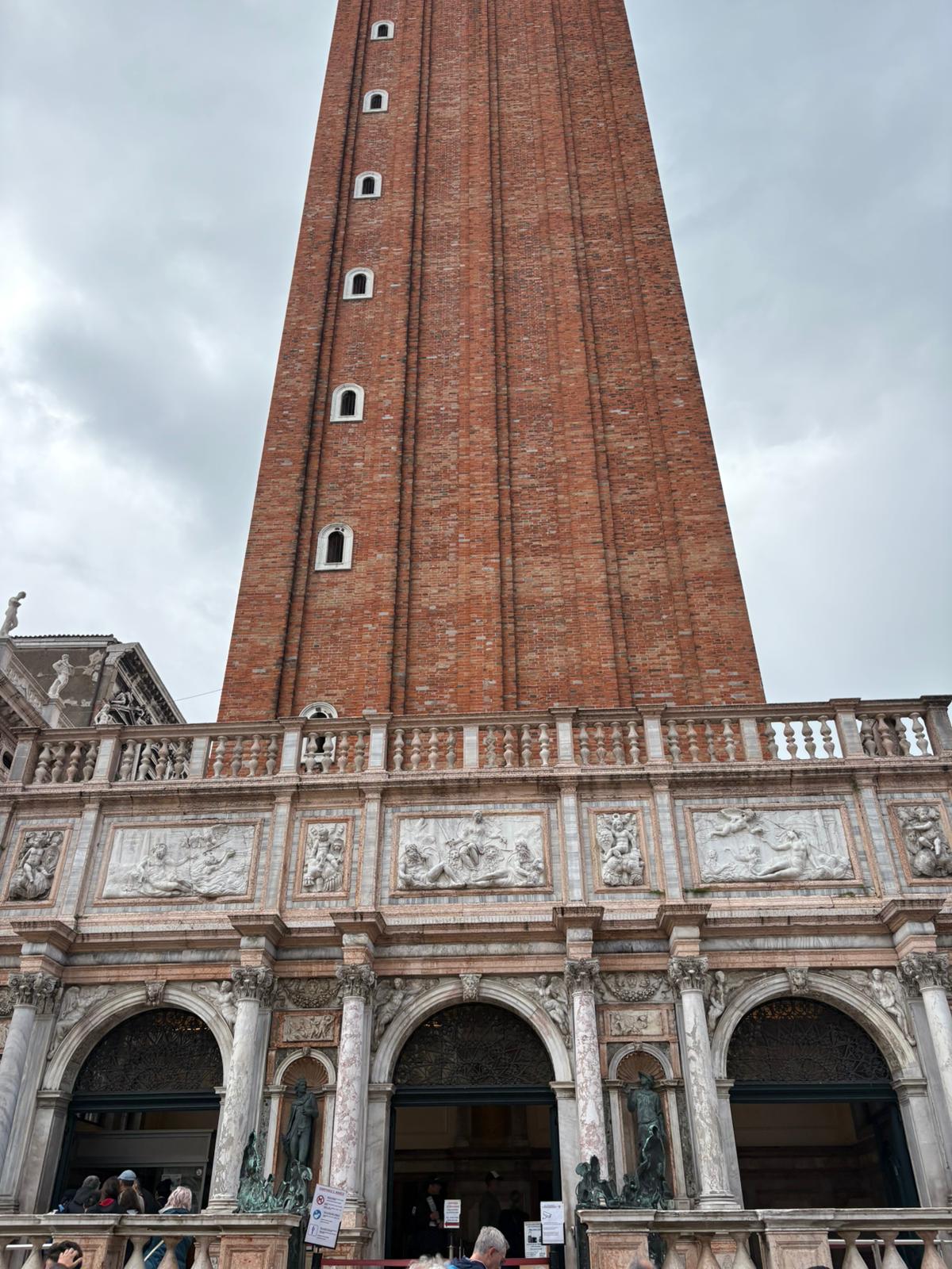 Classical Brick Tower with Sculpted Facade Tall brick tower with arched windows, decorative base with sculptures, overcast sky background.