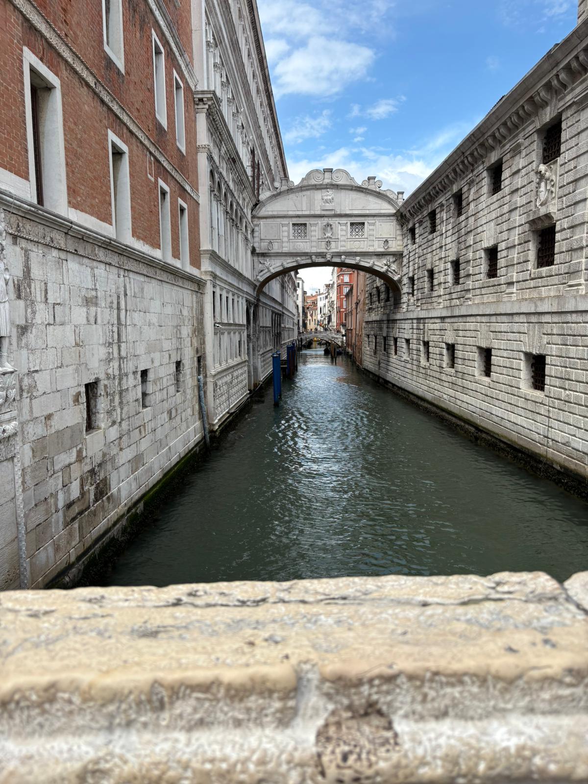 Narrow canal with historic stone buildings and arched bridge under partly cloudy sky.