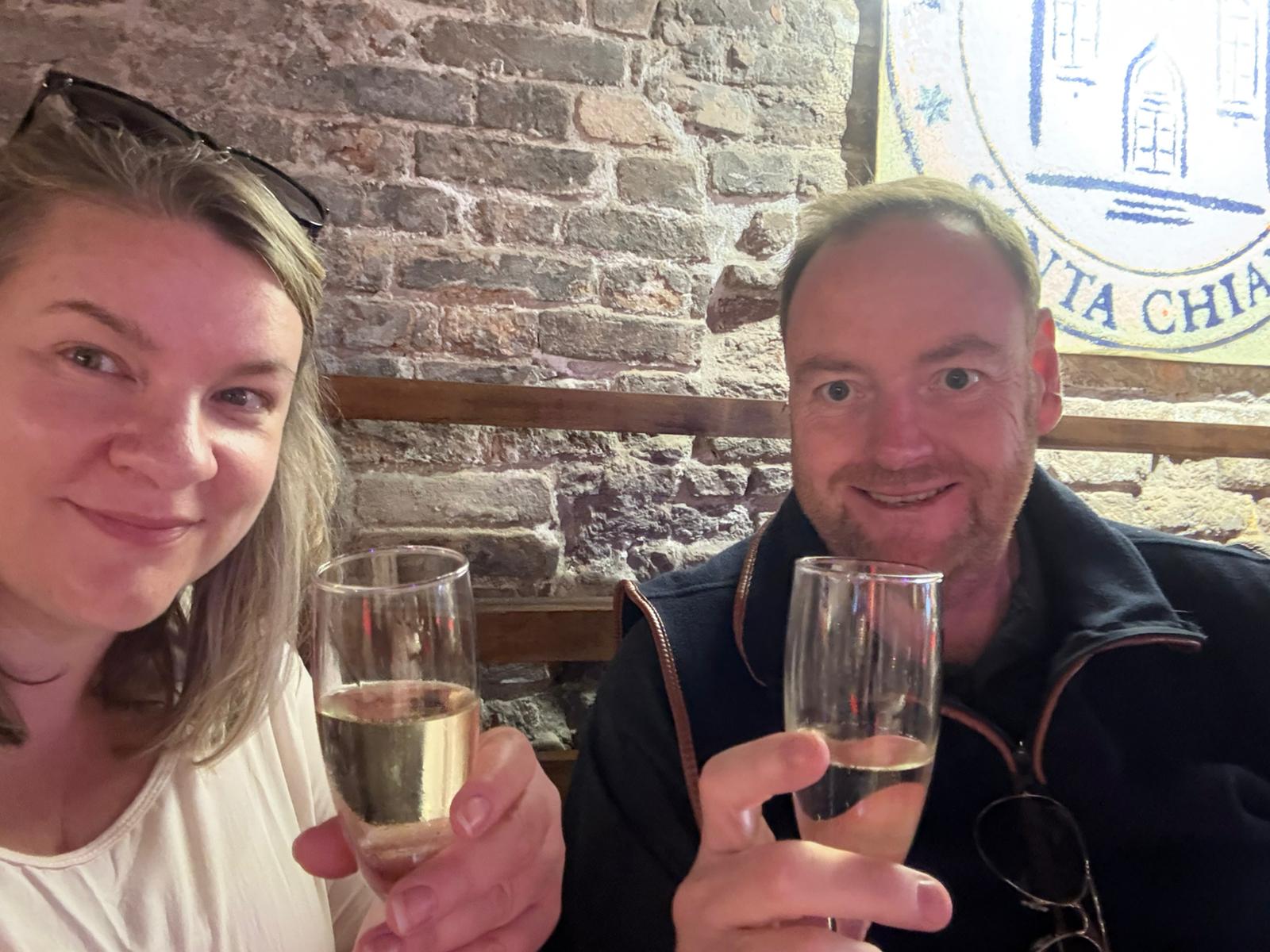Man and woman with champagne, sitting in rustic setting, brick wall, partial logo visible.