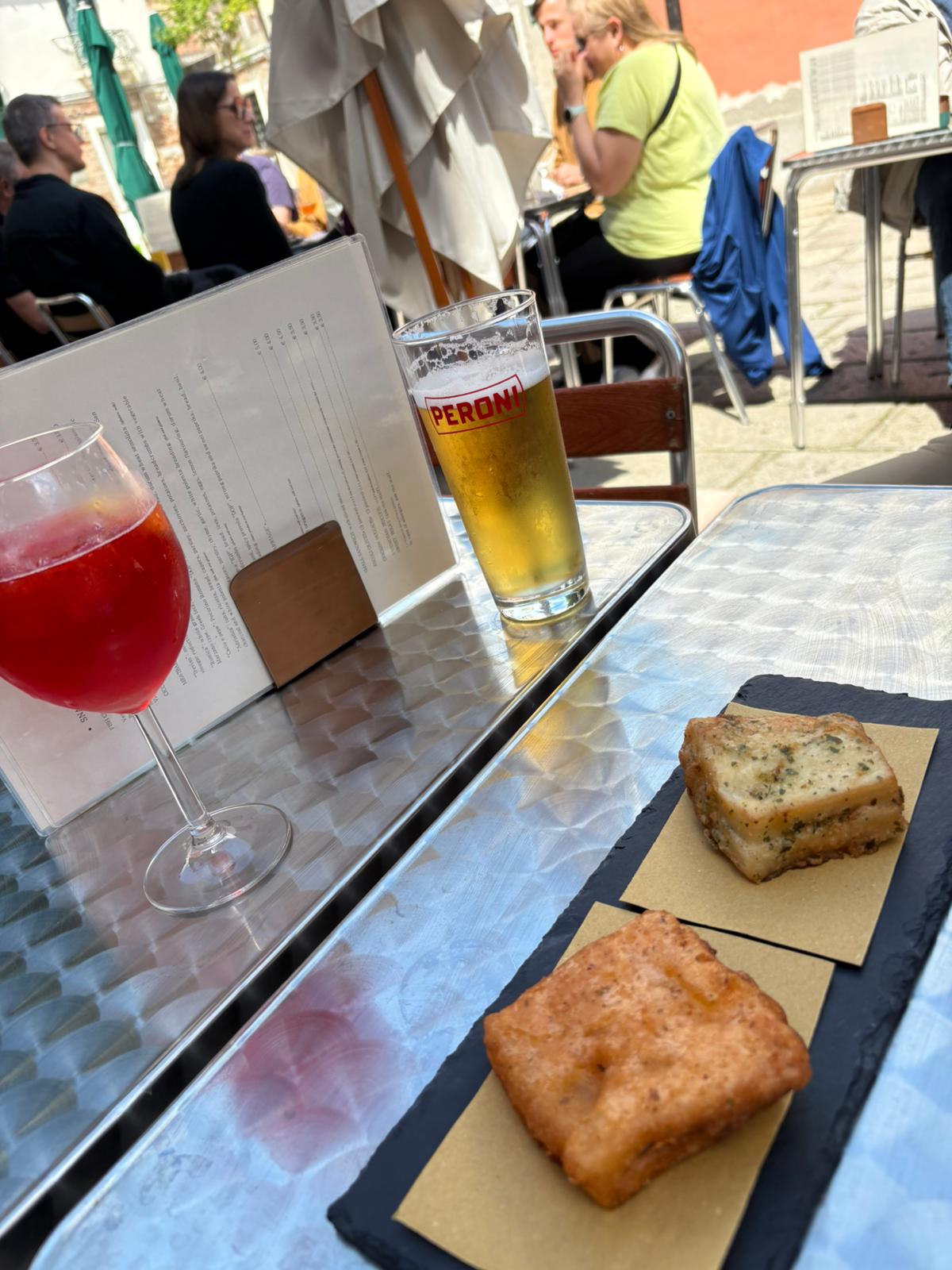 Outdoor café table with Peroni beer, red cocktail, fried food; people enjoying open-air setting.
