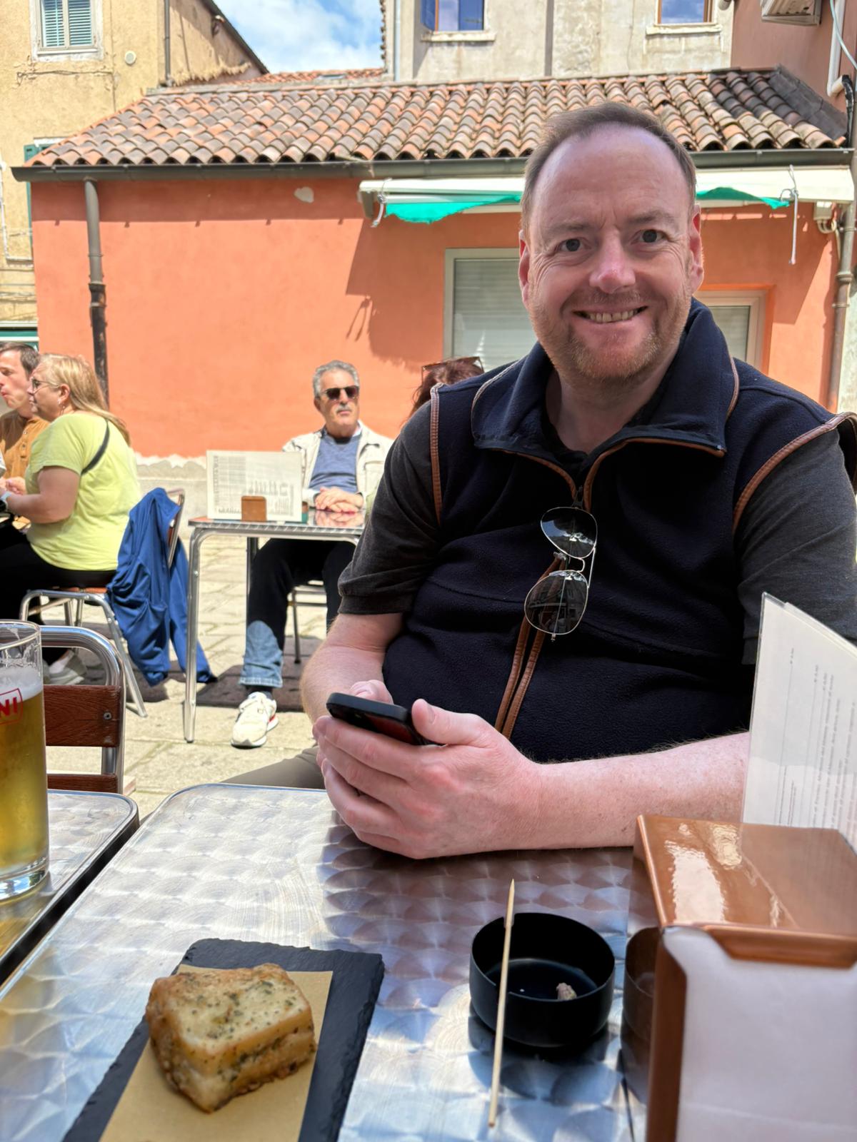 Outdoor Cafe Moment with Smartphone Person smiling at outdoor cafe, holding phone, with food, beer, and others in background.