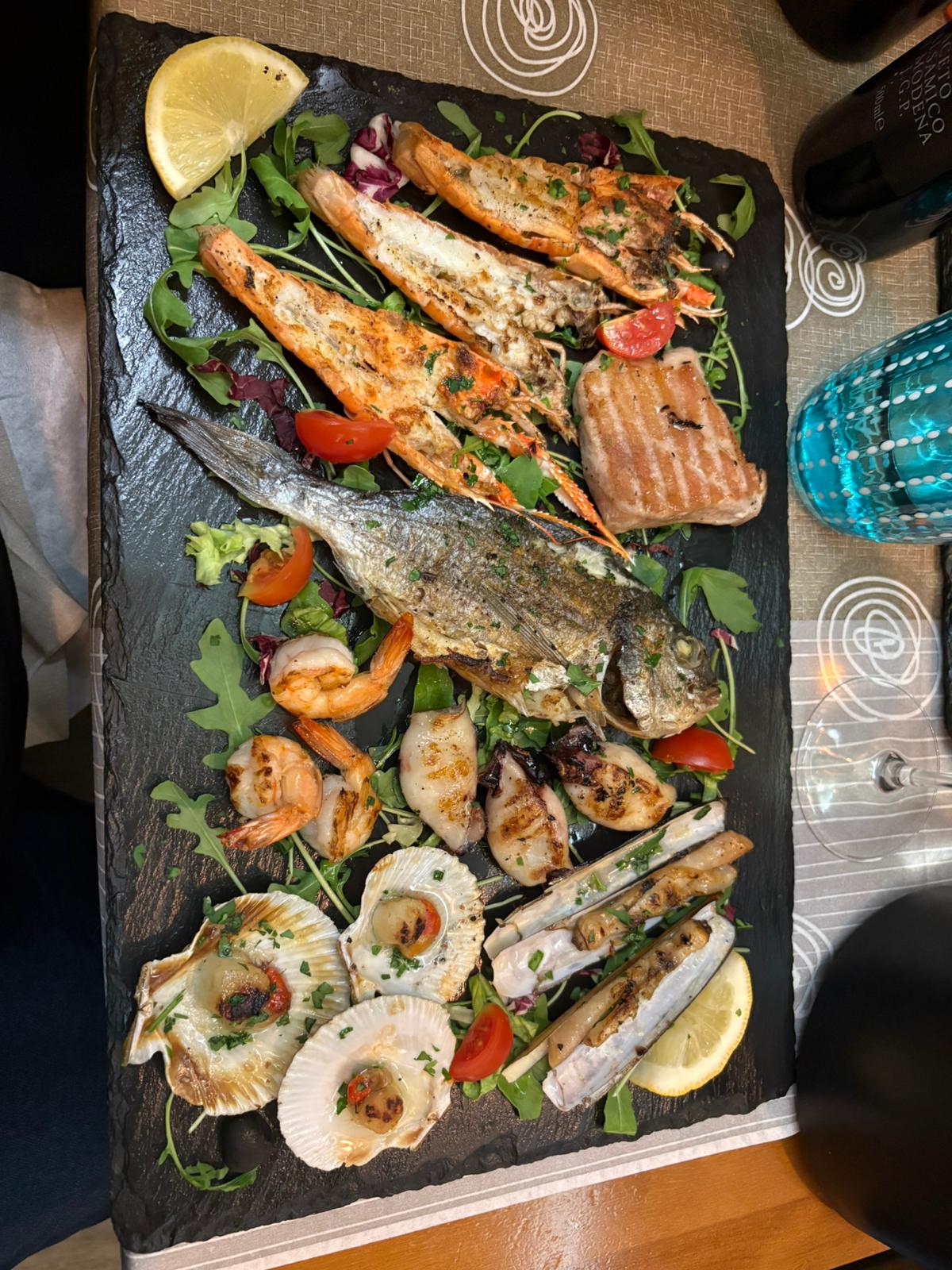 Assorted seafood on slate platter with grilled fish, prawns, scallops, squid, greens, lemon wedges.
