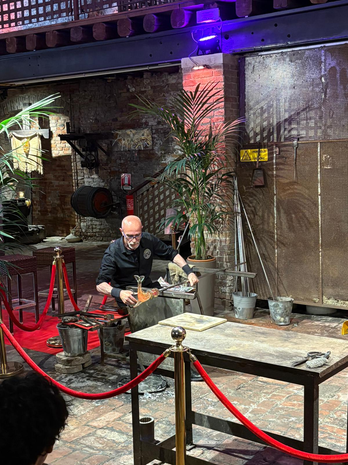 Glassblowing Artistry in Industrial Setting Person performing glassblowing in an industrial setting with red ropes and potted plants nearby.