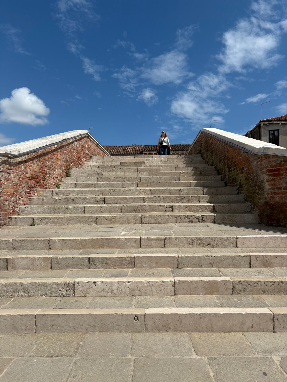 Person sitting atop stone stairs with brick walls, under clear sky with scattered clouds.