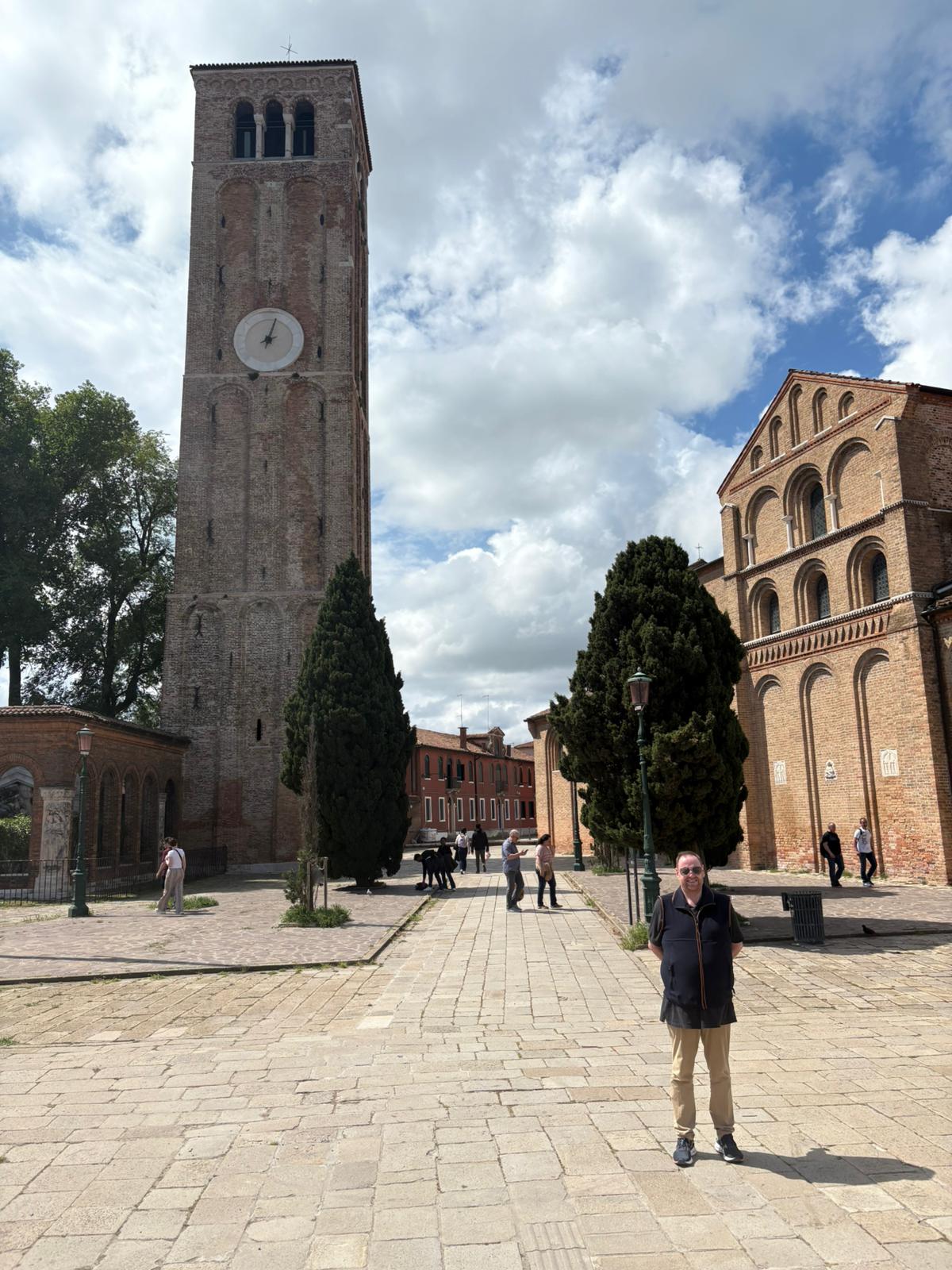Tall brick bell tower with clock, arched window building, people in sunny courtyard.