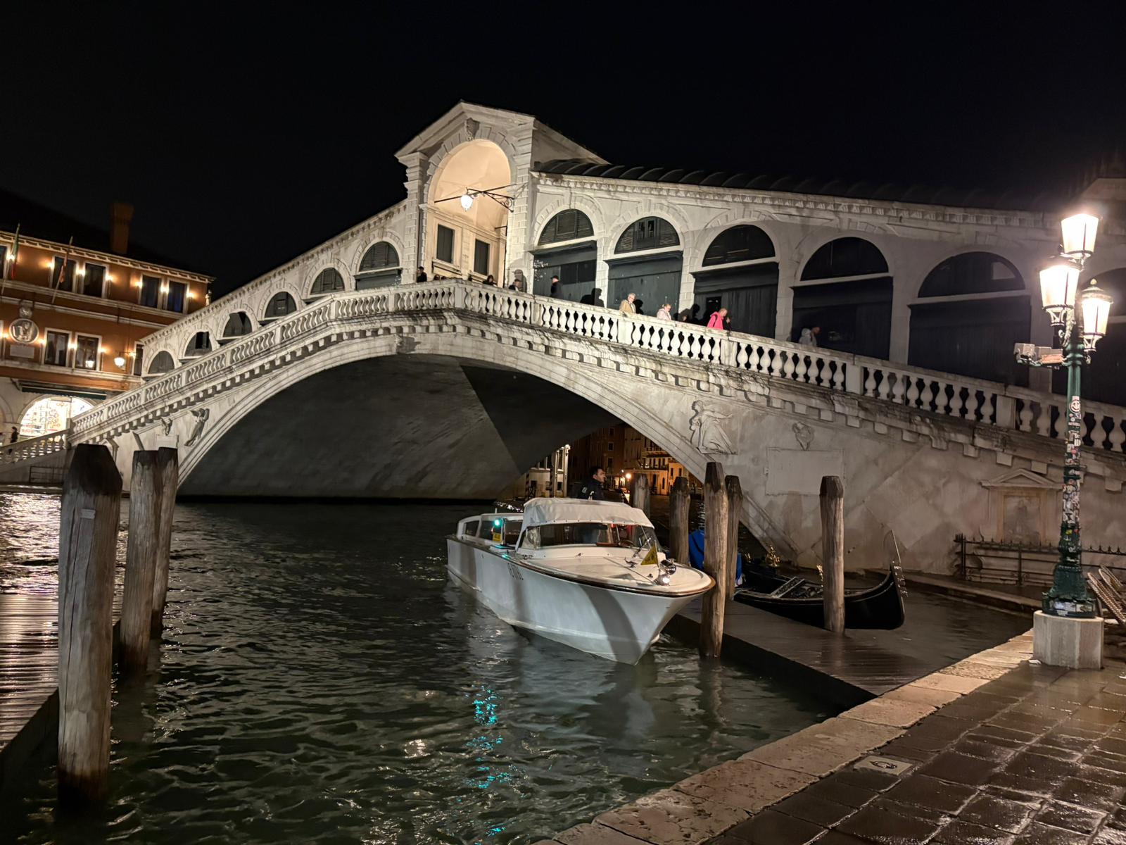 Stone bridge over canal at night with boat and gondolas, illuminated by nearby building lights.