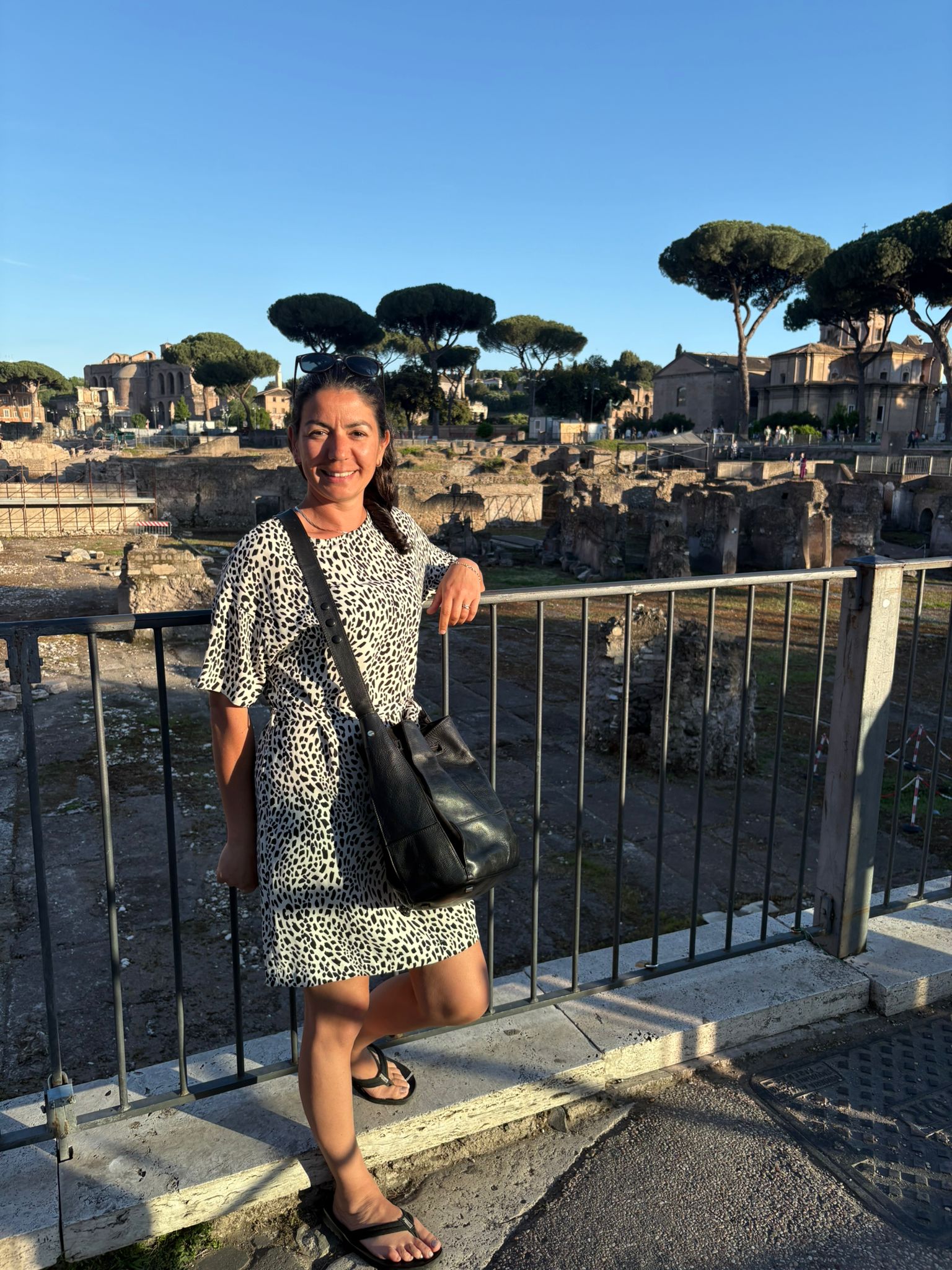 Person in patterned dress stands by railing with historic ruins and trees in sunny background.